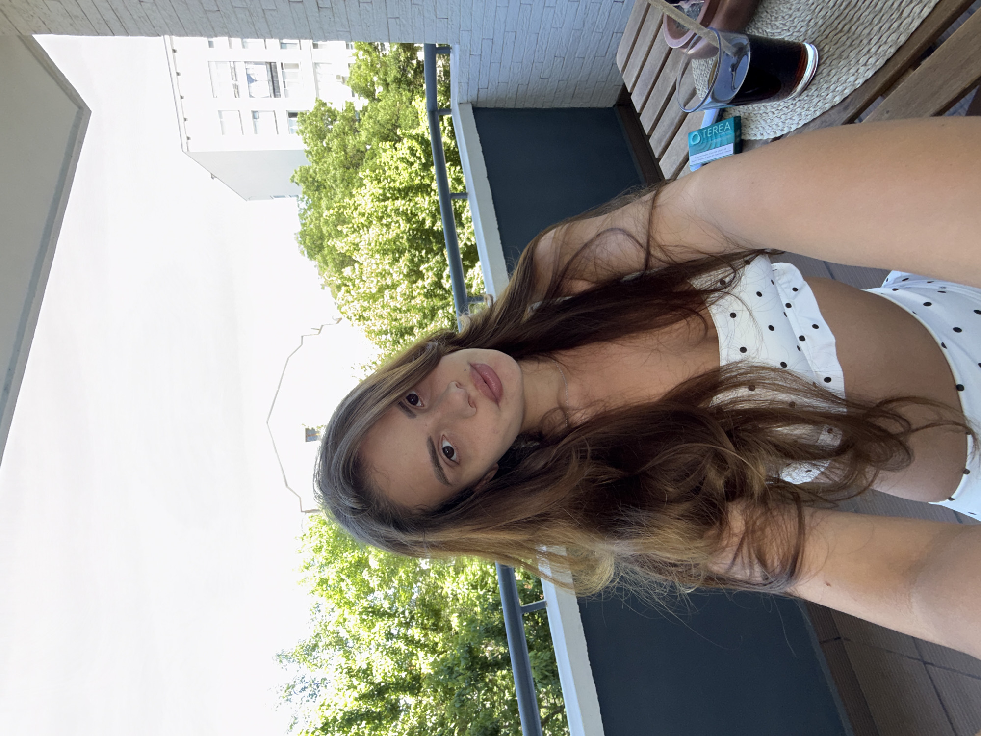 A young woman with long, brown hair is taking a selfie on a balcony. She is wearing a white bikini with black polka dots. She has a light complexion and is looking directly at the camera with a relaxed expression. In the background, there is lush greenery and a white building. On a wooden table next to her, there is a glass of dark liquid with a straw and a pack of TEREA cigarettes. The scene appears to be a residential balcony overlooking a city.