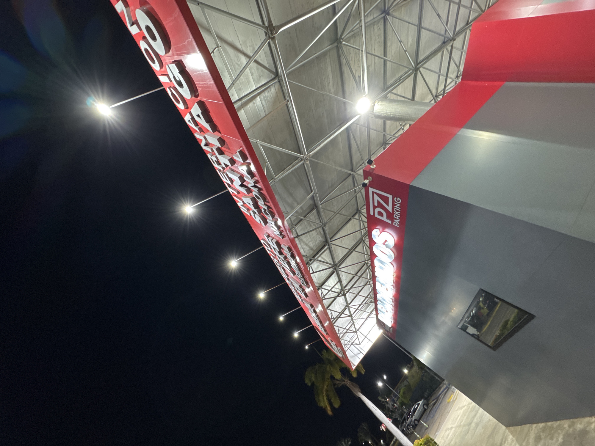The image depicts an exterior view at night, presumably of a parking structure in Porlamar, Venezuela, given the Spanish signage “BIENVENIDOS” (Welcome) and the urban environment. The structure is characterized by a red and gray color scheme and features a metal framework overhead. The signage includes "PZ PARKING" in red and white lettering. Light poles illuminate the scene. A palm tree is visible in the background along with parked vehicles. The scene is angled upwards, creating a distorted perspective.