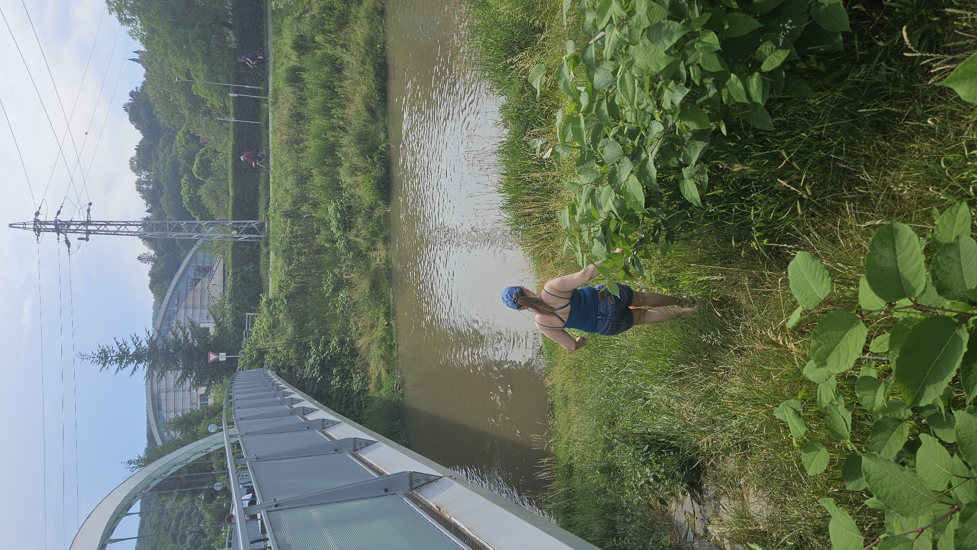 The image depicts a scenic outdoor area with a river, a bridge, and a woman walking towards the water. In the background, there is a modern building with a curved roof, a high-voltage electrical tower, and lush green trees. The woman, wearing a blue hat, a blue tank top, and denim shorts, is walking down a grassy slope towards the river. The river appears murky and calm. The bridge is a pedestrian bridge with a metal railing. There are cyclists visible on a path in the distance, and a traffic sign near the building. The overall setting suggests a park or recreational area within a city. This location is the area near the rehabilitation center and the bridge crossing the Torysa river in Prešov, Slovakia.