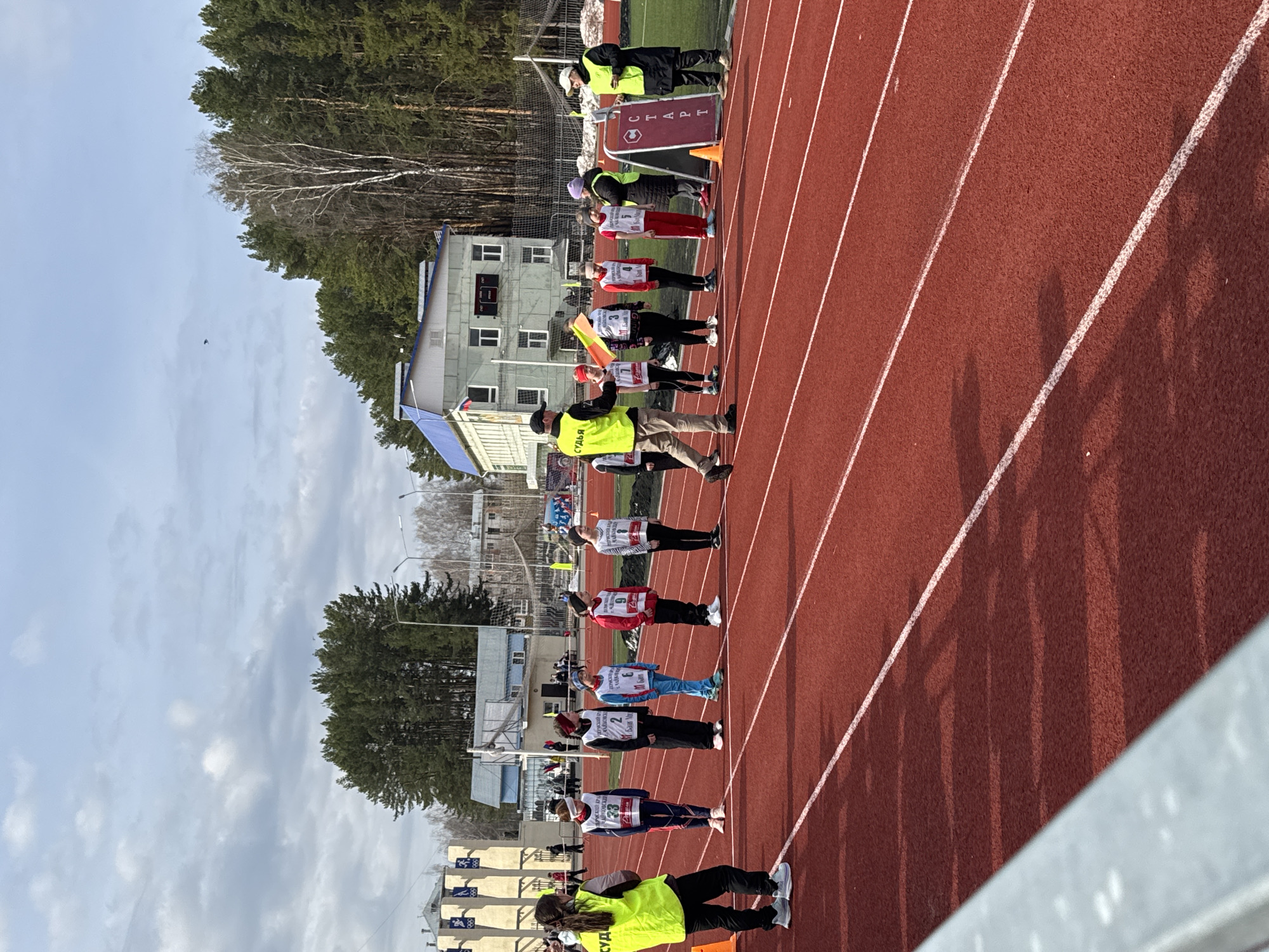 A straight-on outdoor shot shows runners on a track in Chaikovsky, Russia. The runners are lined up on the track, facing away from the camera. They are wearing sports attire and racing bibs with numbers. Adults stand adjacent to the racers and behind the start line. A red banner that reads "START" in Russian can be seen. The track is red and marked with white lines, dividing the lanes. A building with a blue roof is behind the track. Trees are in the background. The sky is cloudy.