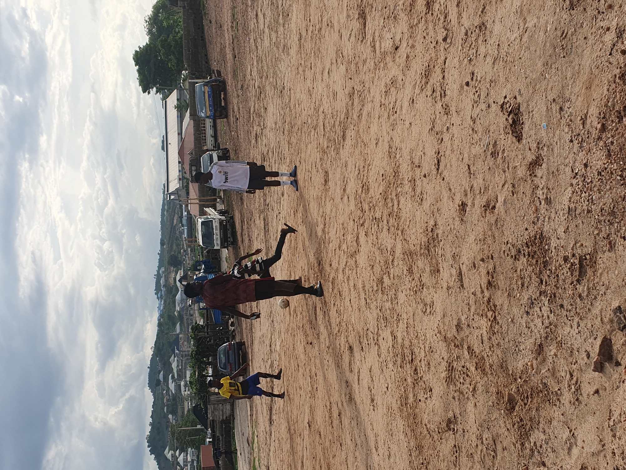 The image captures a dusty open area in Lokoja, Nigeria, where three young men are playing soccer. The ground is sandy and uneven. In the background, a street is lined with parked cars and trucks. Buildings and trees dot the hillside leading to a cloudy sky. One individual is wearing a white jersey, another a yellow shirt and blue shorts, and the third a maroon shirt. The players are actively engaged in a soccer game. A ball is visible.