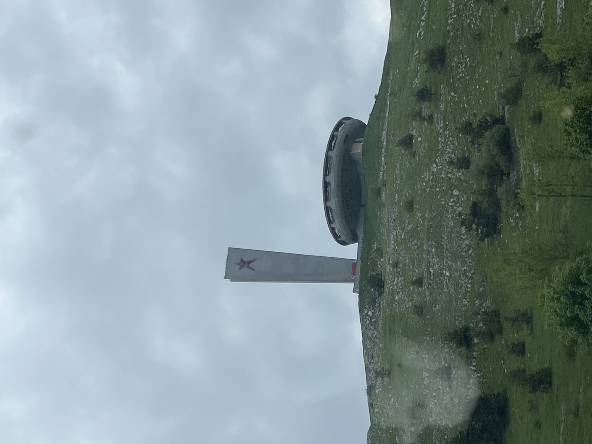 The image shows the Buzludzha Monument in Bulgaria, situated atop a mountain. The monument consists of a disc-shaped structure and a tall pylon with a red star. The surrounding environment is characterized by a grassy mountainside with scattered trees. The sky is overcast. The monument is located near the town of Kran.