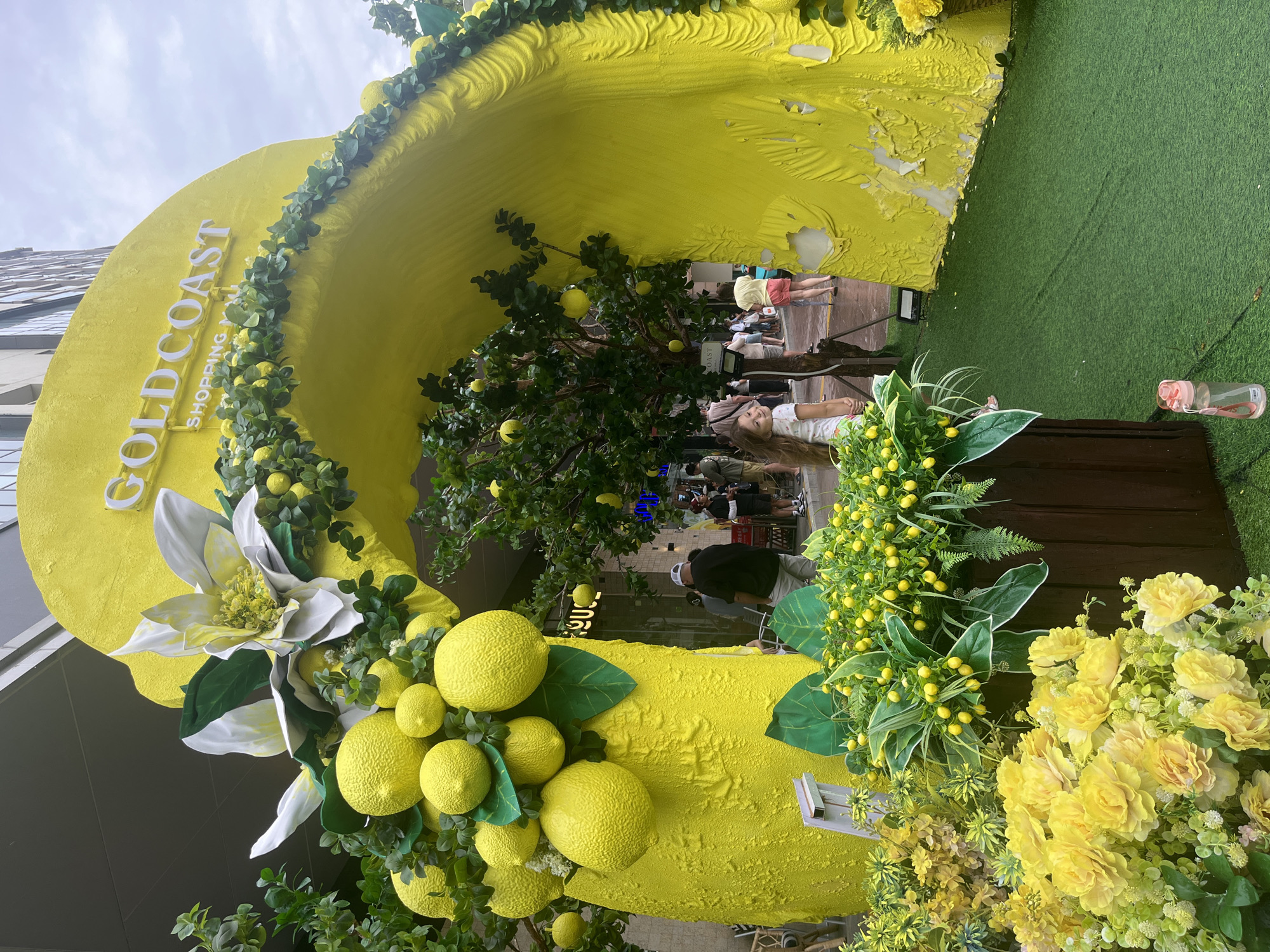 The image shows an outdoor display with a large, yellow structure resembling a halved lemon. "GOLD COAST SHOPPING" is written in white letters on the lemon. The structure is decorated with artificial lemons, green foliage, and a large white flower. A tree with artificial lemons inside the structure is visible. People are walking through the structure. A grassy area is present to the right of the lemon display, and a building is in the background.