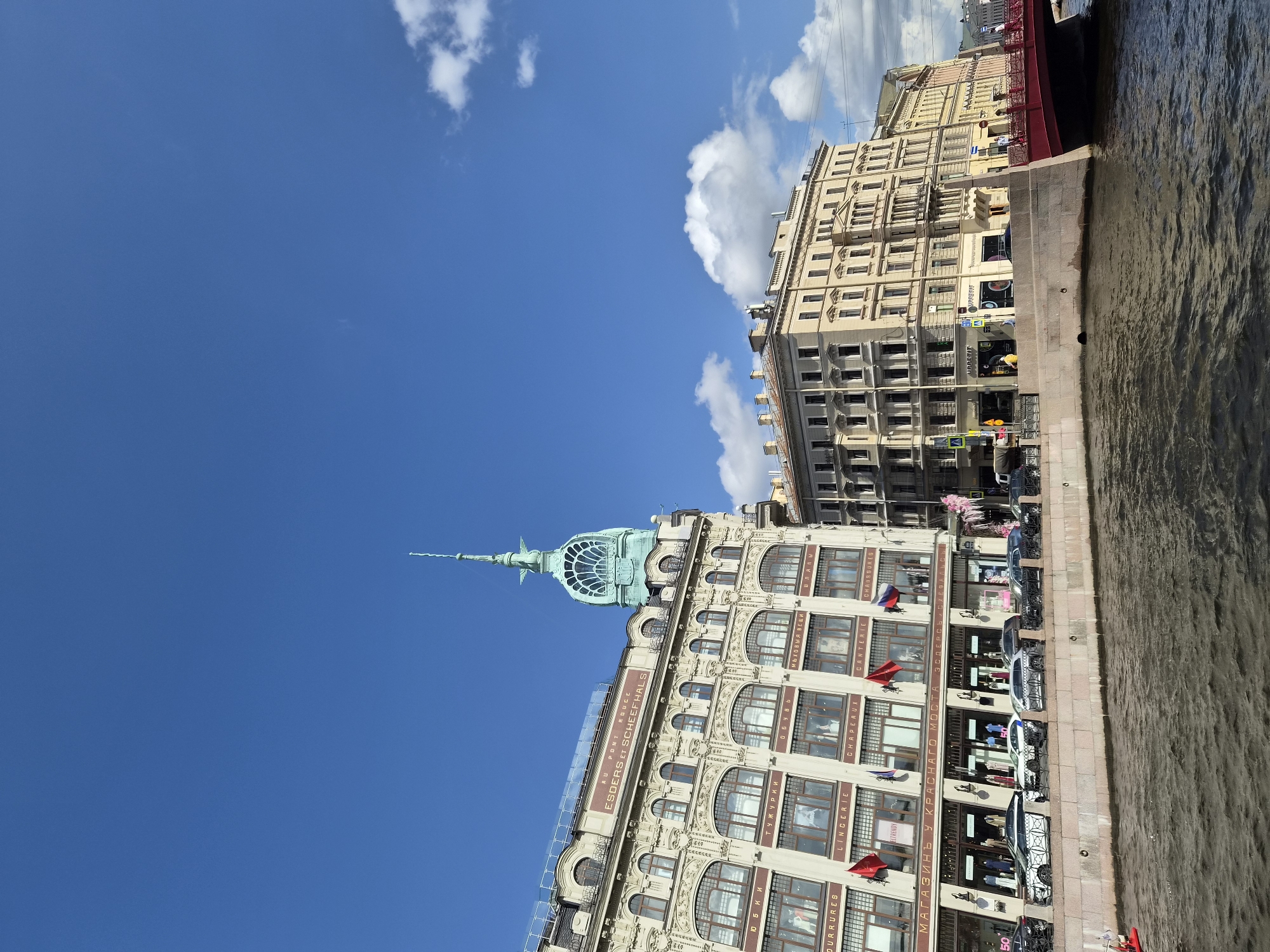 This is a low-angle, eye-level shot of a canal in St. Petersburg, Russia, on a clear day. The buildings on the left and center are ornate and multi-storied, with arched windows and decorative facades. The building on the left features a prominent green spire with a distinctive arched window structure at its base. The building in the center is a lighter color with many windows, and signage in Russian is visible on its lower levels. Several cars are parked along the embankment in front of these buildings. A red bridge spans the canal in the right side of the frame. The canal water is dark and choppy, reflecting the sky and buildings. Scattered white clouds dot the bright blue sky. No people are clearly visible.