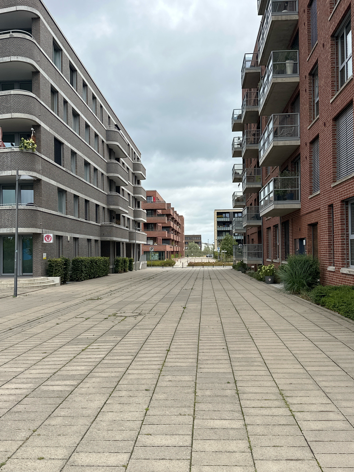The image depicts a pedestrian pathway lined with modern apartment buildings on either side. The pathway is paved with light-colored rectangular concrete slabs, with some sparse vegetation growing between them. The building on the left is characterized by its gray brick facade and curved balconies. The building on the right features a red brick facade and glass-enclosed balconies. In the distance, additional apartment buildings with similar architectural styles are visible, suggesting a planned urban development. The sky is overcast with clouds. This scene is located in Bremen, Germany, specifically in the Überseestadt district, an area undergoing significant redevelopment with a focus on modern residential and commercial architecture. There are no people visible in the image, and no specific actions or interactions are taking place. The environment is a clean, open urban space.