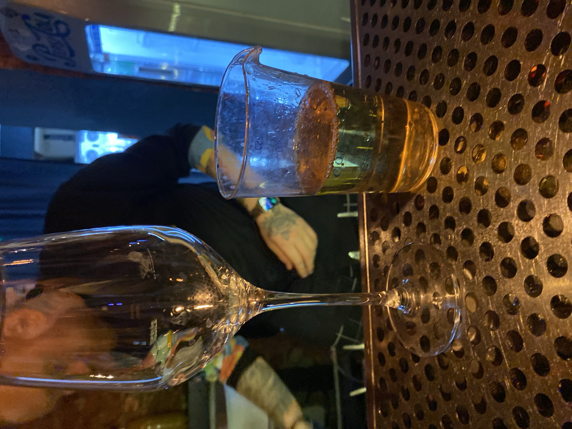 The image shows a bar setting, potentially in Izhevsk, Russia. Two glasses are present: a wine glass filled with a clear liquid and a lab beaker containing a yellow beverage with visible bubbles. The wine glass is positioned towards the lower left corner, while the beaker is located in the upper right. In the background, an individual with tattoos on their arm is visible. The surface beneath the glassware appears to be a perforated metal bar top.