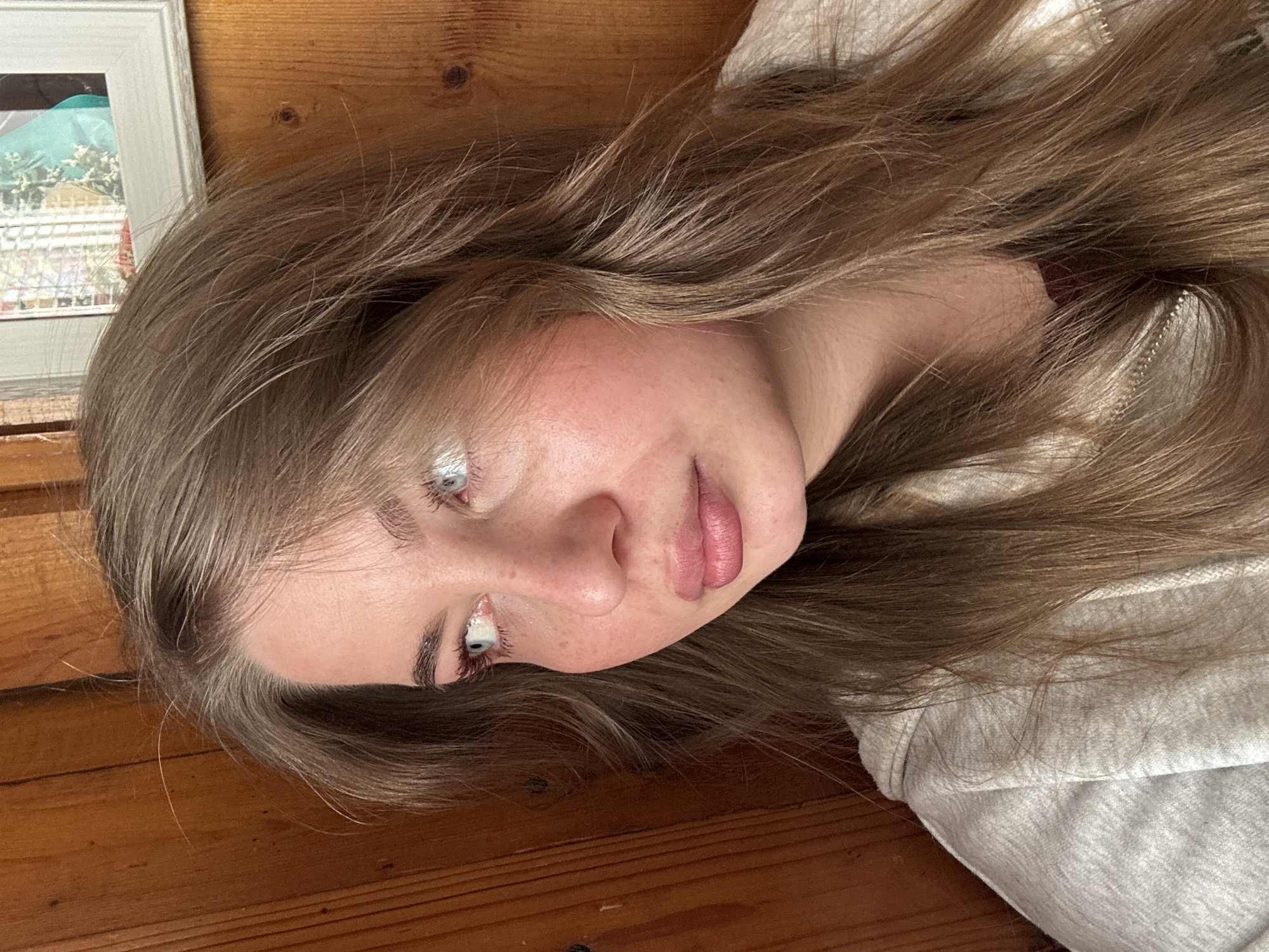 A close-up, portrait-style shot captures a young person, likely female, lying down on a textured wooden surface. The individual has long, wavy light brown hair, which frames the face and spreads across the wooden background. Their light blue or grey eyes are a focal point, featuring distinct white or light-colored eyeliner or glitter on the lower lash line and inner corner, which creates a striking contrast. Freckles are visible on the bridge of the nose and cheeks, and the lips are full with a natural, slightly pinkish hue.
The person is wearing a light grey hooded garment, possibly a sweatshirt or hoodie, part of which is visible on the right side of the frame. The background consists of horizontal wooden planks with visible grain and knots, suggesting an indoor setting with natural wood paneling or flooring. In the upper left corner, a portion of a light-colored picture frame is visible, enclosing what appears to be a landscape or cityscape scene with buildings and greenery. The overall mood is relaxed and contemplative, with the subject looking slightly away from the camera. There is no visible text in the image. Given the provided context, the location is Уральское, Russia.
