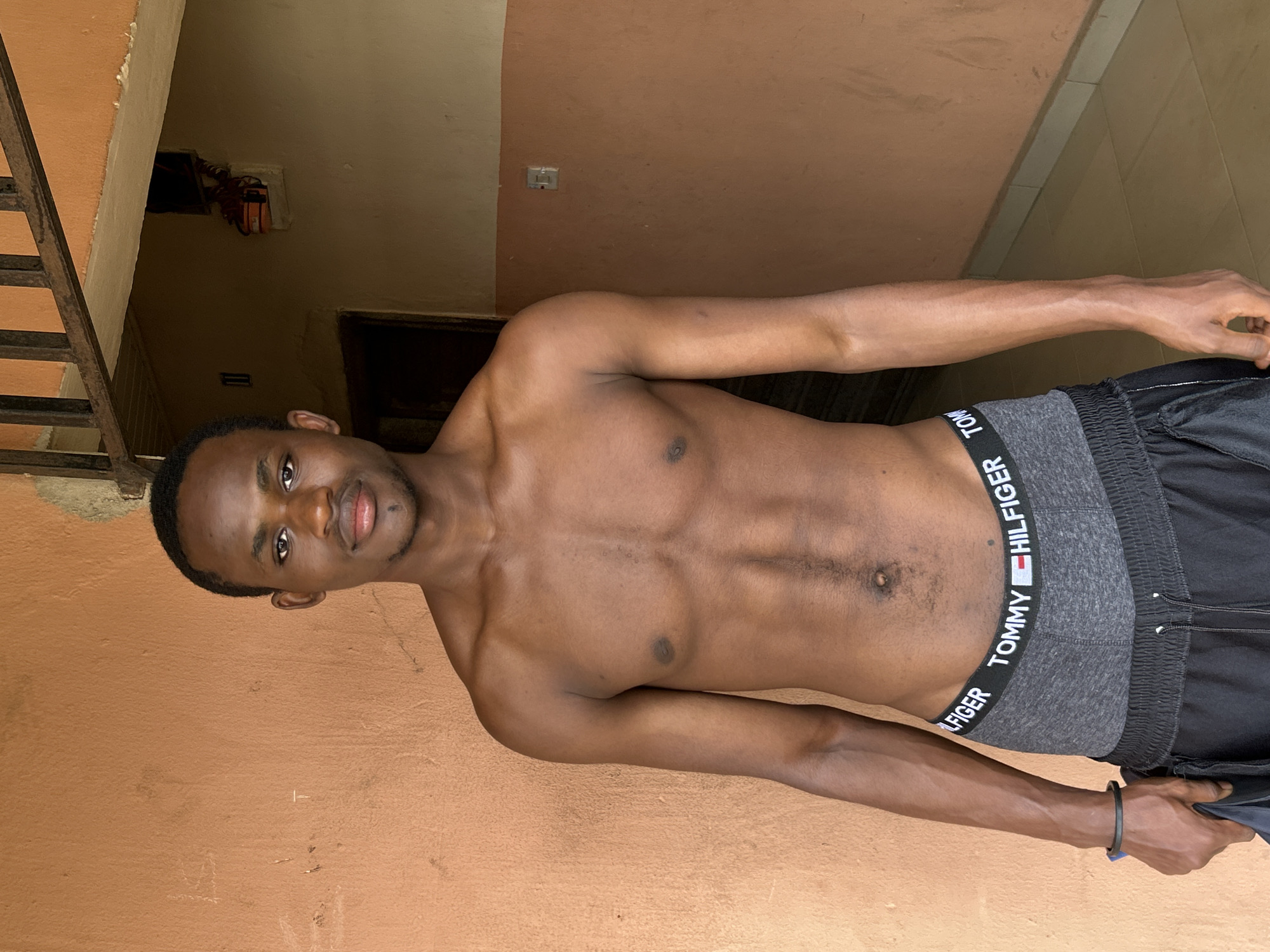 A shirtless young man with dark skin stands against a textured, peach-colored wall. He is wearing gray Tommy Hilfiger boxer briefs and dark shorts. His body is turned slightly to the side, and his gaze is directed towards the viewer. His facial expression is neutral. In the background, there is a glimpse of a dark interior space with visible electrical wiring and a light fixture. A metal railing is visible on the left side of the image. The lighting suggests it is daytime, but no specific weather conditions are apparent. The presence of the textured wall and the doorway leading into a dimly lit interior could indicate an outdoor or semi-outdoor setting, possibly a residential area.