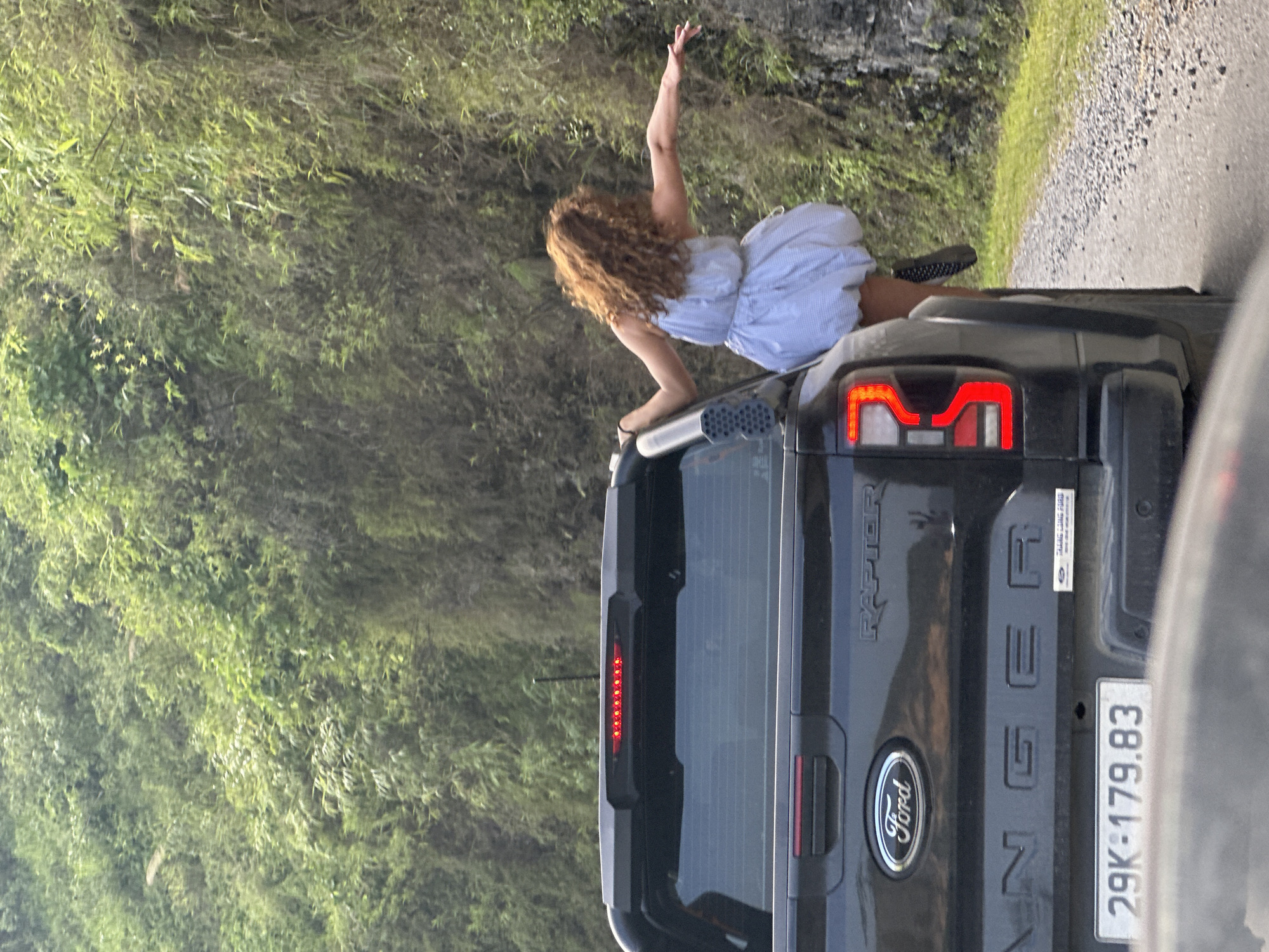 A woman with curly hair, wearing a light-colored dress and black shoes, is on the back of a black Ford Ranger pickup truck. She is leaning over the edge with one arm raised. The vehicle's license plate reads "29K-179.83." The environment appears to be rural, with a dirt road and dense green vegetation on the hillside. The location is Du Già Commune, Vietnam.