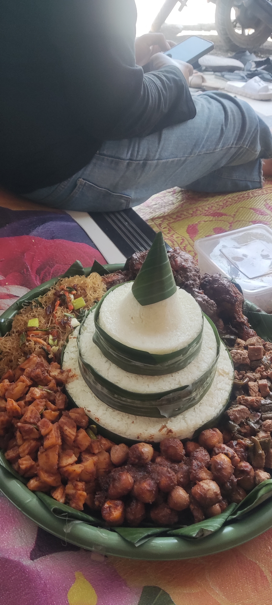 In Gebang, Indonesia, a tiered rice cone, known as nasi tumpeng, is prominently displayed on a green platter lined with banana leaves. The tumpeng is surrounded by various side dishes, including fried noodles, cubed fried tofu, and seasoned chickpeas. In the background, a person is seated, wearing a dark long-sleeved shirt and blue jeans, and is engrossed in using a mobile phone. The setting appears to be outdoors, possibly a yard or a street, with a motorcycle visible in the distance and a patterned mat on the ground.