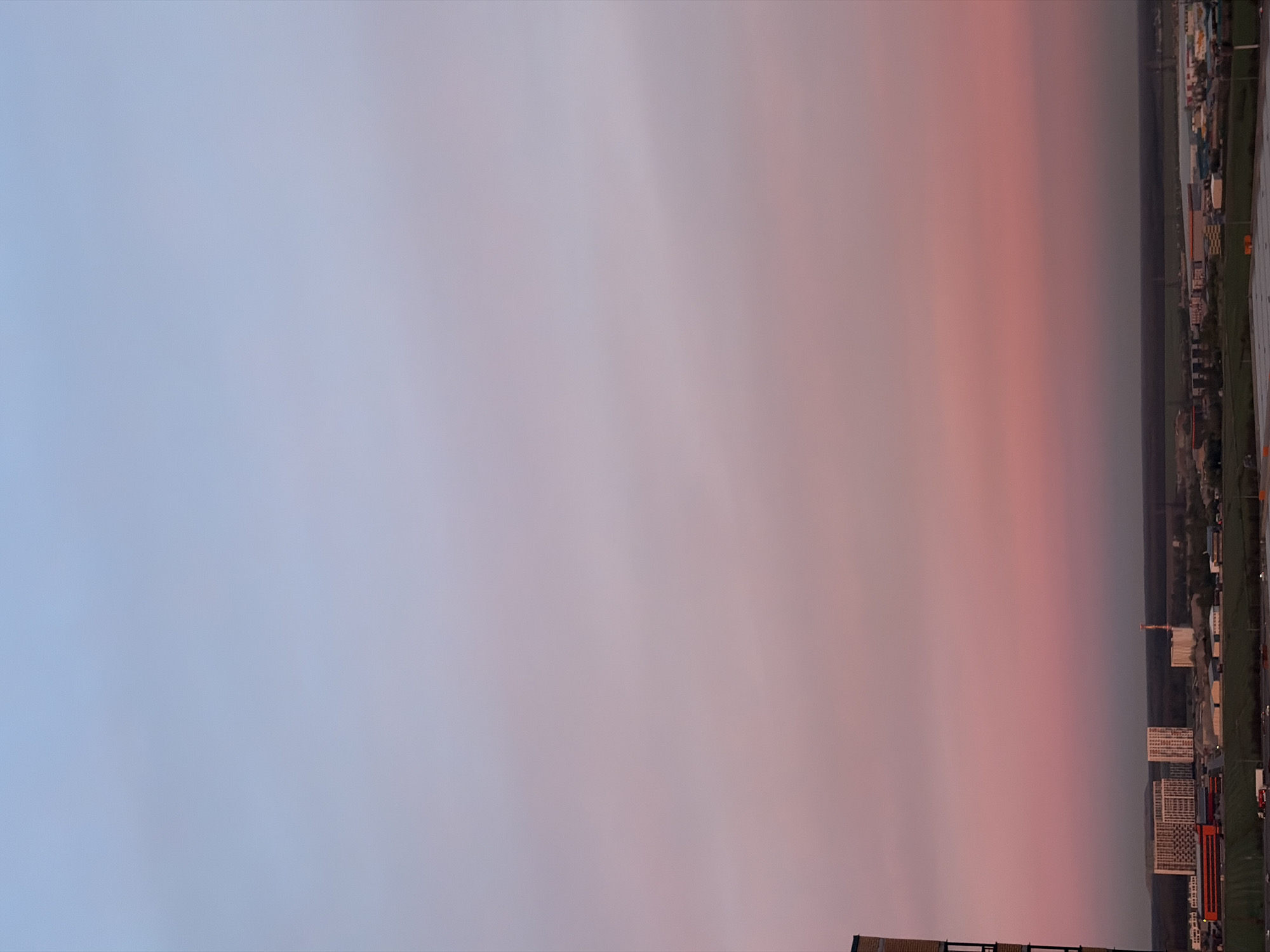 The image depicts a sunset over what appears to be a cityscape. The sky transitions from a light blue on the left to a pinkish-orange hue on the right. At the bottom right of the frame, there are buildings, possibly residential complexes, interspersed with green areas and infrastructure. The general appearance of the buildings and overall layout suggest the scene may be in Saint Petersburg, Russia. There are no people visible in the frame. The primary subject is the sunset and the adjacent cityscape.
