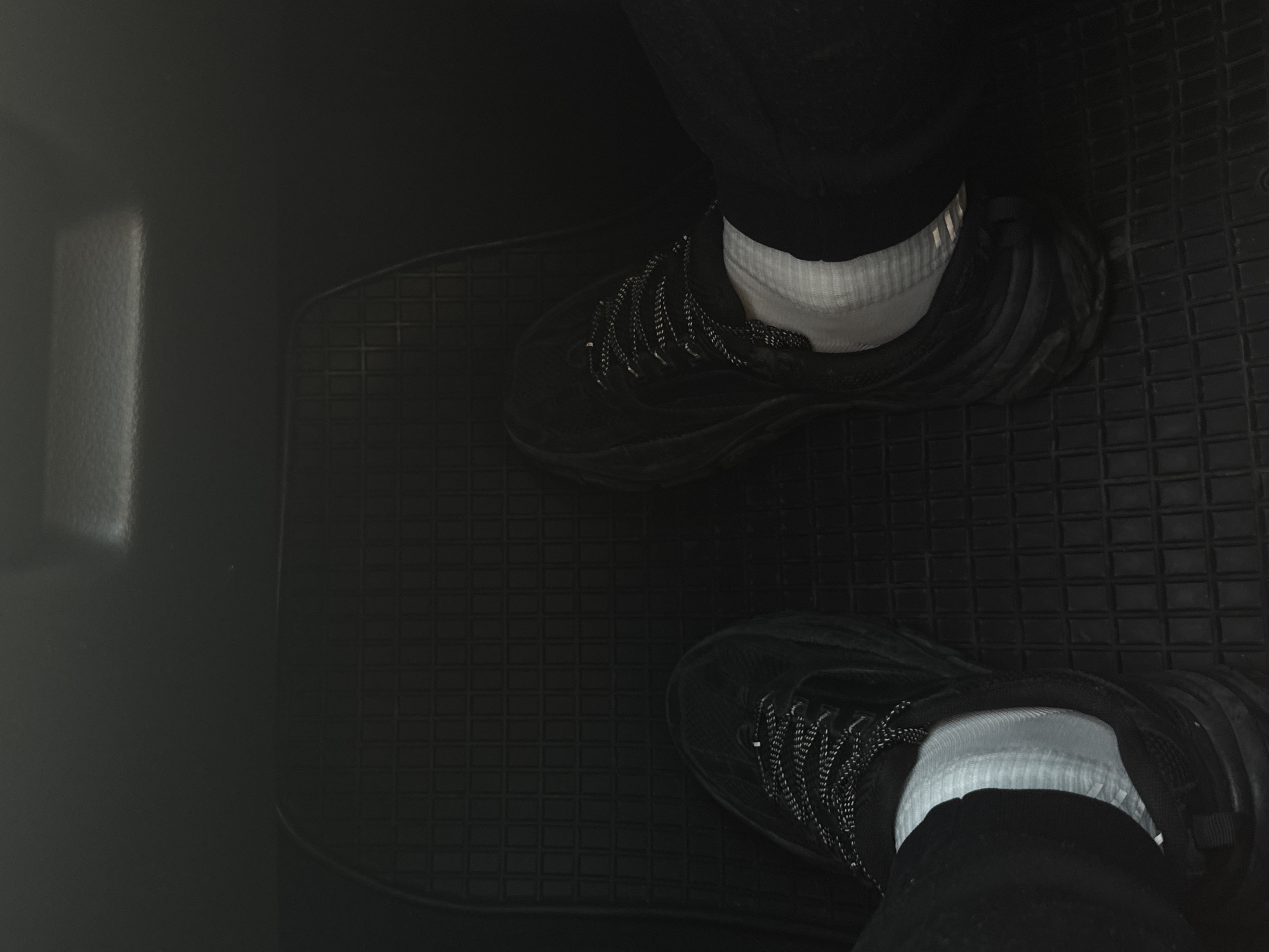 A top-down indoor shot depicts a pair of black athletic shoes being worn with white socks and black pants in the footwell of a vehicle. The footwell is lined with a black rubber mat with a square grid pattern. To the left is a smooth, dark gray or black surface, possibly the car's interior. The location is unknown, and there is no mention of Diadovo, Bulgaria, in the image.