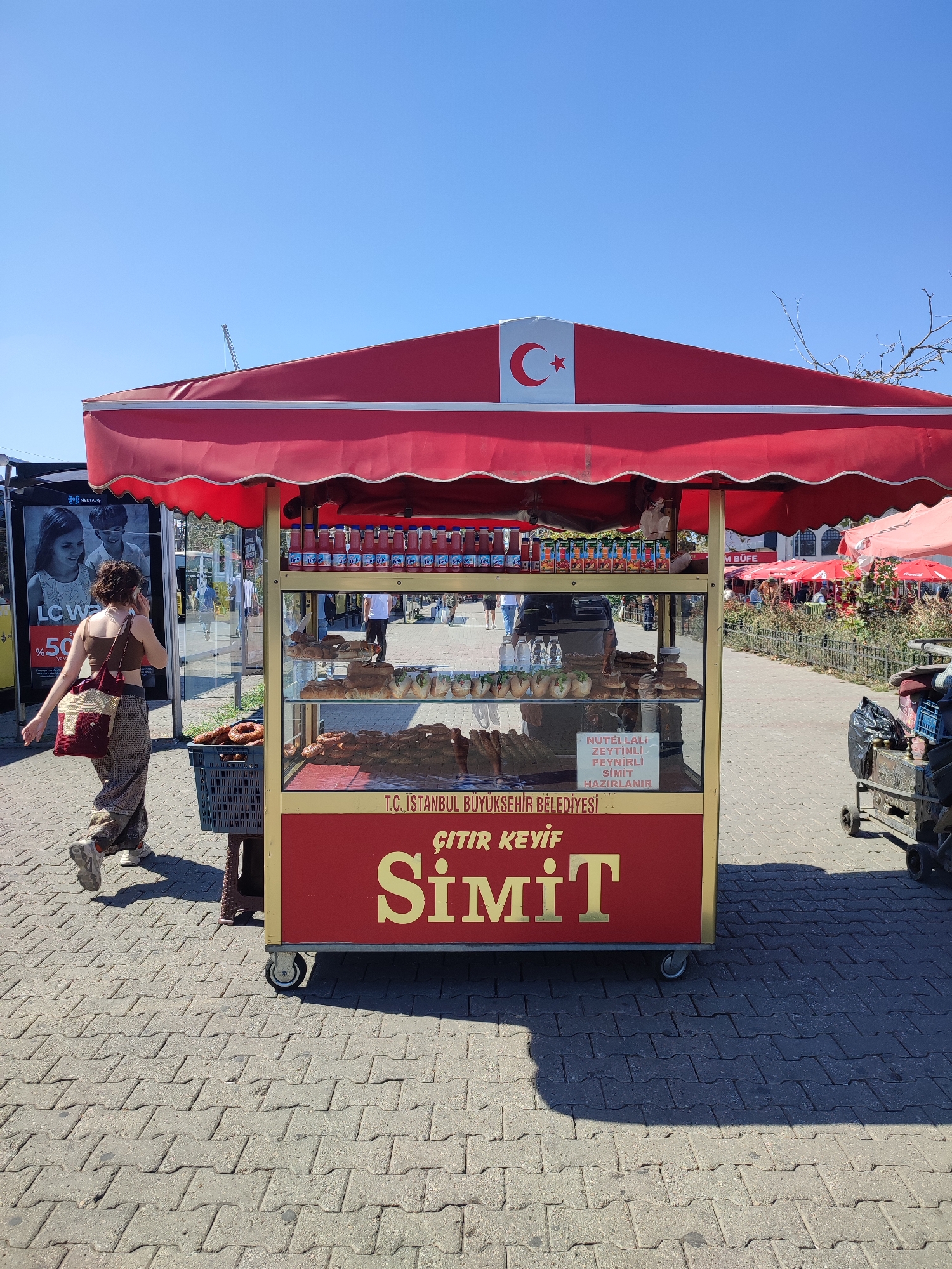 A red and gold food cart, displaying baked goods and bottled drinks, is parked on a cobblestone sidewalk in Kadıköy, Turkey, under a clear blue sky. The cart is branded with the Turkish flag and text indicating "T.C. İSTANBUL BÜYÜKŞEHİR BELEDİYESİ" (Republic of Turkey, Istanbul Metropolitan Municipality). A woman with brown curly hair, wearing a brown crop top and patterned pants, walks past the cart, talking on her phone and carrying a bag over her shoulder. Behind the cart, several red umbrellas are visible over outdoor seating areas, suggesting a public space with food vendors. The environment appears to be an urban outdoor setting, with some greenery and other people in the background.