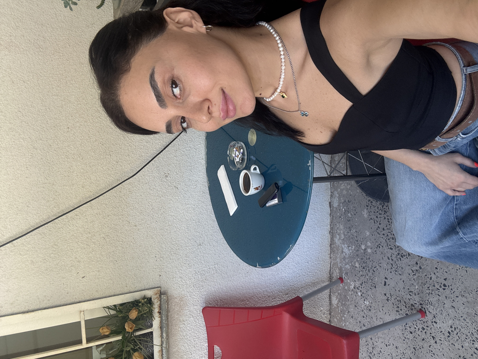 A woman with dark hair and light skin is sitting at a small, round, teal table. She is wearing a black, criss-cross top and blue jeans. She has two necklaces on, one with pearls and a gold pendant, and another with a silver cross pendant. She is also wearing small hoop earrings. In front of her on the table is a white cup of coffee, a cigarette lighter, and an ashtray filled with cigarette butts. To her left is a red plastic chair with metal legs. Behind the table is a white stucco wall with a window containing yellow tulips. The ground is a textured grey concrete. The image appears to be taken outdoors in a cafe or patio setting.