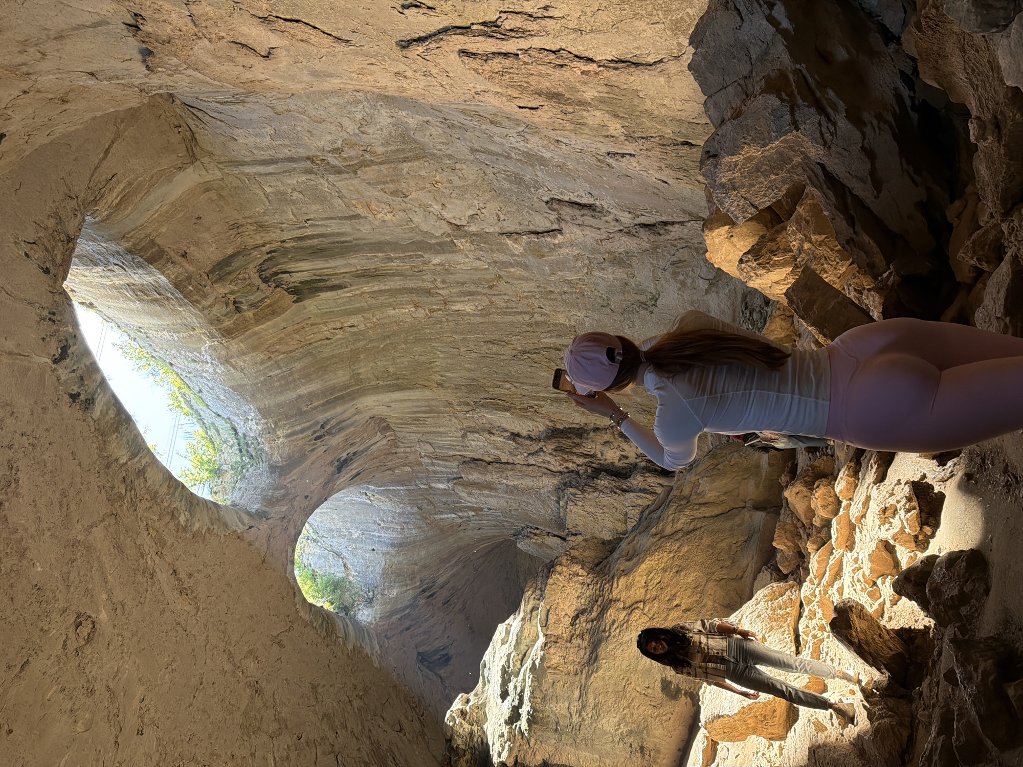 Two women are standing in Devetashka Cave, located near Karlukovo, Bulgaria. One woman in pink leggings and a white shirt is holding up her cell phone to take a picture. Another woman in a flannel and jeans is standing in the lower right. The cave has two prominent holes in the ceiling that let in light.