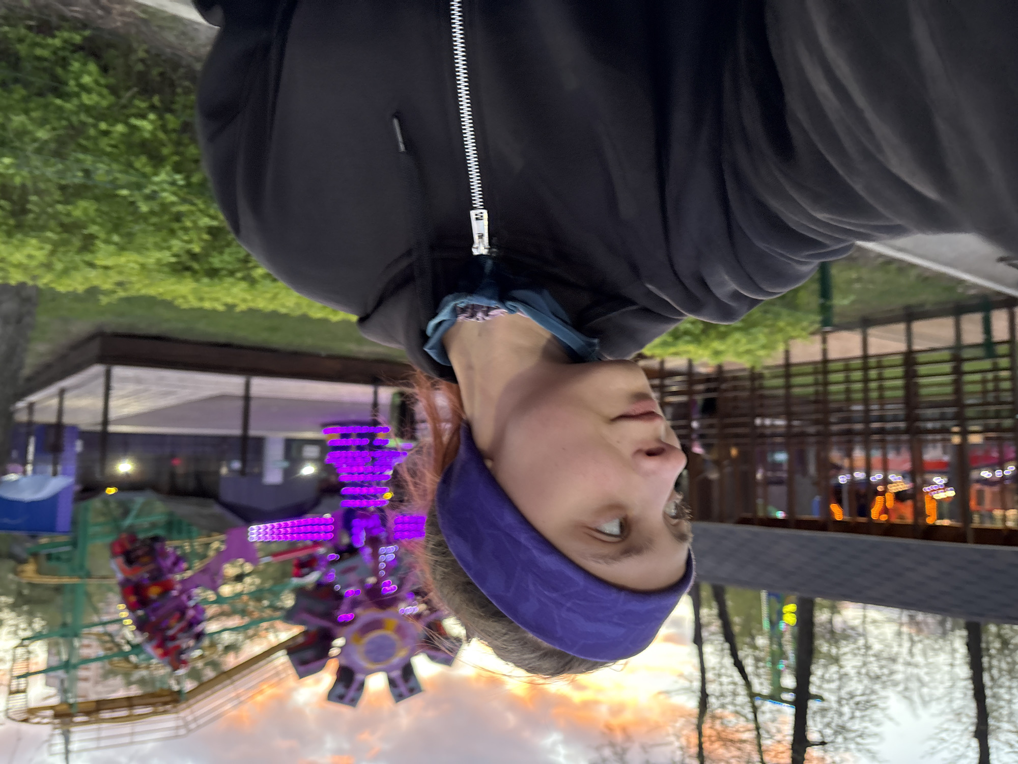 A photograph shows a person wearing a navy blue headband and a black hooded jacket with a zipper. In the background, there's an amusement park ride with purple lights. The surrounding environment shows trees and a structure that looks like a roof or awning. The city of Perm, Russia, is not evident from the image.