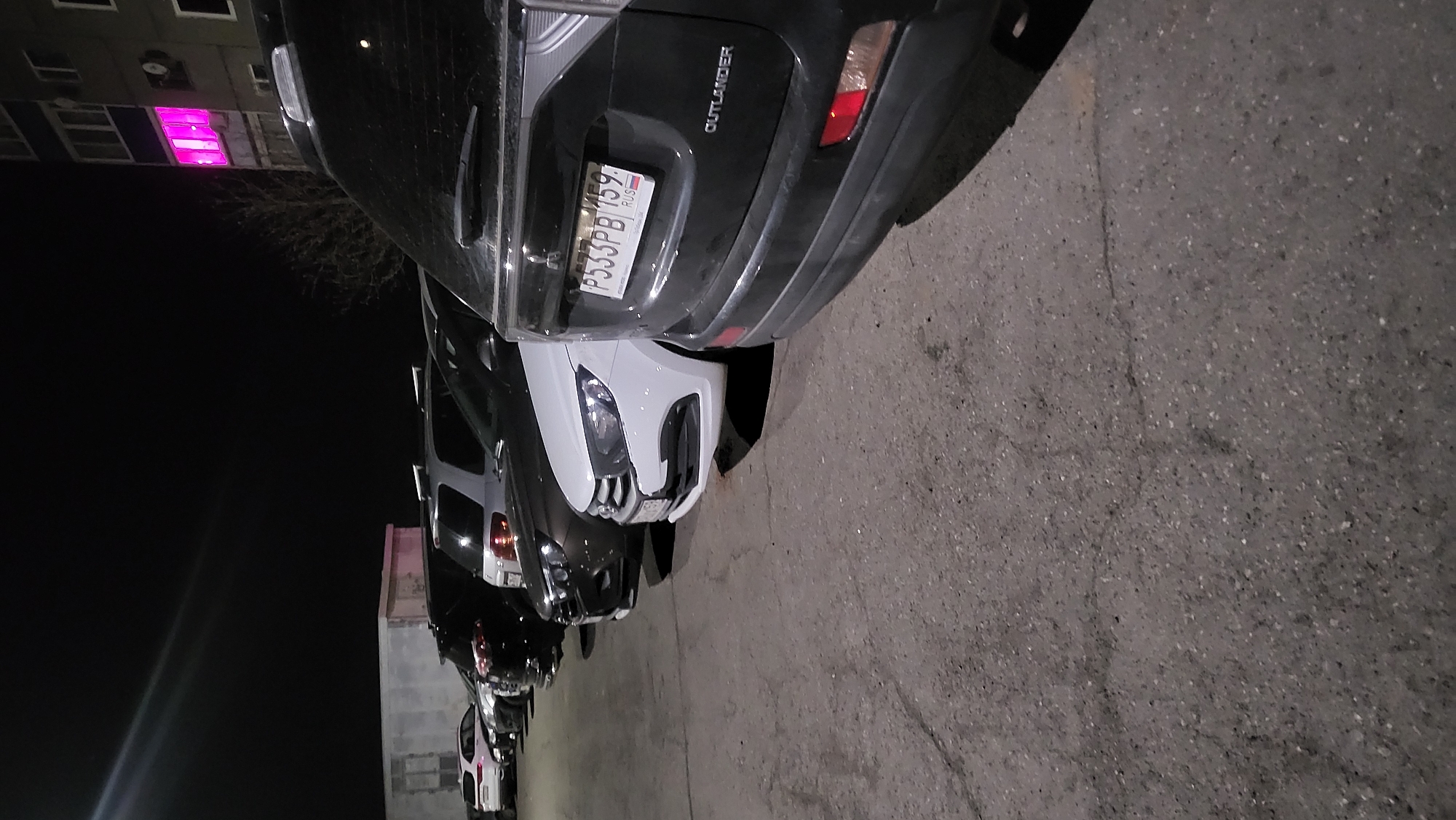 The image shows a row of parked cars at night. The first car is a dark-colored Mitsubishi Outlander, identifiable by the "OUTLANDER" badge. Its license plate reads "P533PB 159 RUS." Behind it is a white car, followed by a series of other vehicles parked in a row. The scene is located in the city of Марковский, Russia. A building with at least one window illuminated in pink is visible in the background. The ground is asphalt.