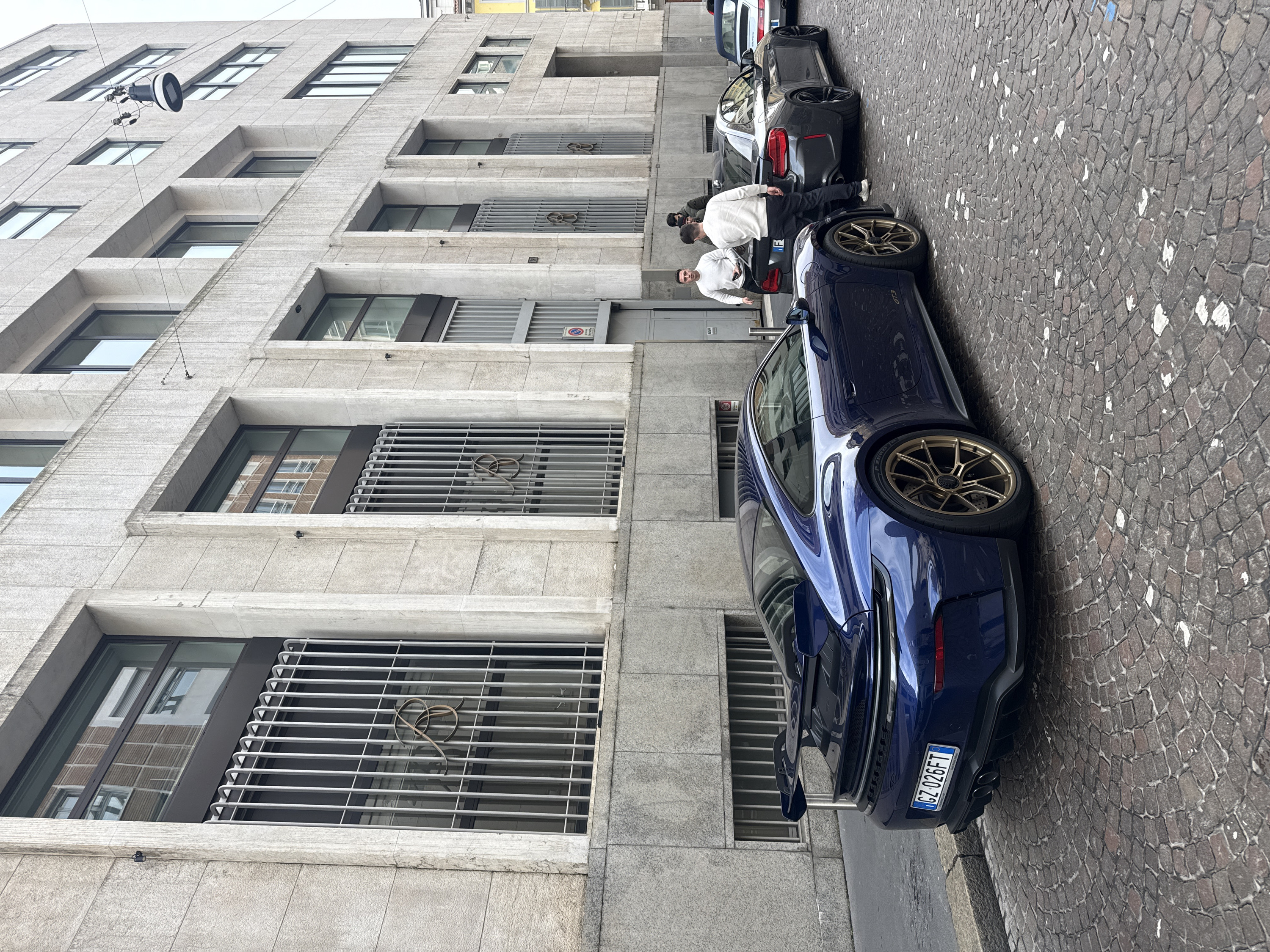 This image depicts an urban street scene in Milan, Italy, on what appears to be an overcast day. The foreground features a cobblestone street, predominantly dark grey with interspersed lighter grey and white patches, suggesting wear or old markings.
Three high-performance vehicles are parked along the left side of the street, adjacent to a raised curb. The closest vehicle, angled away from the viewer, is a dark blue Porsche 911 GT3 (992 generation) with prominent gold/bronze multi-spoke wheels and a large rear wing. Its Italian license plate reads "GZ 026FT." Directly behind it is a dark grey BMW M2 (F87 generation). Further down the street, partially obscured, is a light blue Porsche 911 convertible, likely an older generation.
On the right side of the street, a multi-story building dominates the background. It features a light-colored, textured stone facade with prominent rectangular windows framed in dark material. The ground-floor windows are protected by vertical metal grates, two of which display a distinctive stylized, intertwined "JB" monogram in a metallic finish. A red circular no-parking sign with Italian text "Passo Carrabile" (driveway/access) is visible on the building's facade near an entrance. Several smaller windows are visible on the upper floors. A cylindrical street lamp with a dark base and a wide, flat light source is suspended by wires above the street, connected to the building.
Four adult males are visible near the parked cars. Two men are positioned next to the rear of the dark grey BMW M2; one wears a white sweater and black trousers, while the other wears a dark jacket. A third man in a white sweater and dark trousers stands to the left of these two, holding a mobile phone. A fourth man, partially obscured, wears a dark jacket and is behind the group near the BMW. They appear to be standing and conversing or observing the vehicles.
