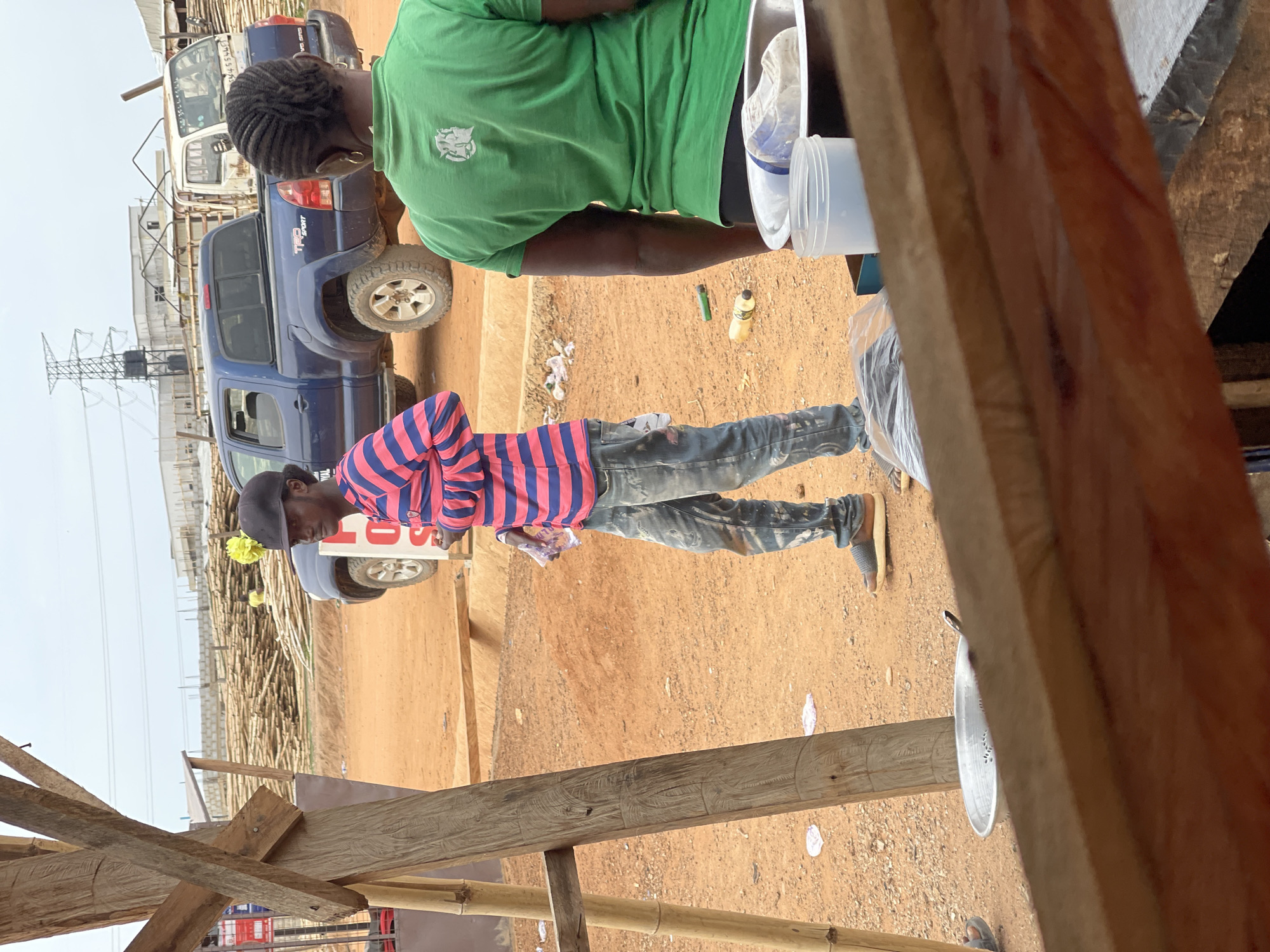 The image captures a scene in what appears to be a commercial area in Gosa, Nigeria. Two individuals are prominently featured. One is wearing a bright green t-shirt and seems to be engaged in some activity involving white containers and a clear plastic bag. The other individual is wearing a horizontally striped pink and dark blue shirt, along with jeans and a dark cap, and is holding a small clear plastic bag, possibly containing food or snacks.
Behind them, two pickup trucks are visible, one white and one blue. In the background, there is a large pile of bamboo stalks, and power lines are seen stretching across the sky. A partially constructed wooden structure is in the foreground, obscuring some of the view. The ground is dirt, and various discarded items are scattered around, including a bottle and a few pieces of trash.