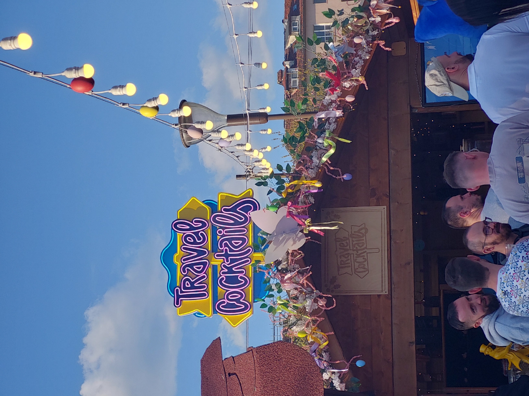 This image, originally rotated 90 degrees clockwise, depicts an outdoor scene at what appears to be a festive market or event during the late afternoon or early evening. The sky is a vibrant blue with sparse white clouds, contrasting with the warm glow of numerous string lights.
A prominent neon sign, brightly illuminated, reads "Travel Cocktails" in a stylized purple font against a yellow outline and blue background. Below and to its right, a wooden stall or structure is heavily adorned with festive decorations, including garlands of green leaves, delicate white and pink flowers, colorful ribbons (pink, yellow, purple), and small pastel-colored ornaments that resemble Easter eggs. A large white paper butterfly is also part of the decor. On the wooden structure, another sign is visible, also bearing the text "Travel Cocktails" in a carved or painted style.
Several men are gathered near the right side of the image, facing towards the wooden stall as if at an outdoor bar. They are casually dressed. One man wears a light blue shirt and a baseball cap, looking upwards. Another wears a grey t-shirt featuring Japanese characters "福冈県" (Fukuoka Prefecture). Their expressions are generally neutral and engaged, suggesting a relaxed social atmosphere.
The illuminated signs and string lights, while the sky is still bright, indicate either dusk or a bright but perhaps overcast day where artificial lighting adds to the ambiance. Part of a large, brown, hat-shaped structure is visible in the bottom-left corner. The overall setting suggests a pleasant, lively outdoor gathering focused around refreshments.