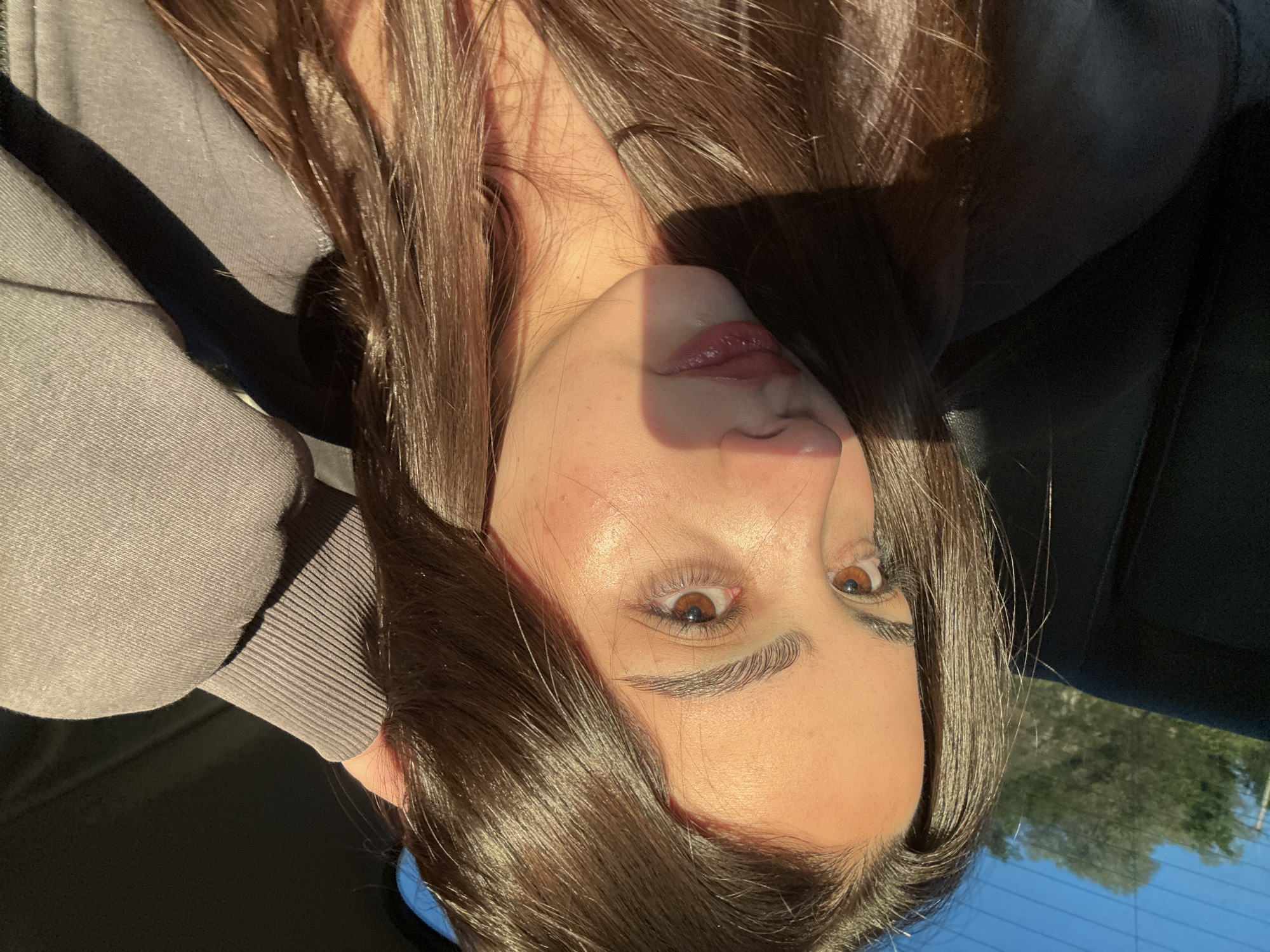 A young woman with brown hair is seated in what appears to be a car. The image is taken from a slightly angled perspective, showcasing her face, particularly her eyes and lips. She is wearing a gray hoodie or sweatshirt. Sunlight is casting shadows across her face and hair. Through the car window in the background, the sky and some greenery are visible. It is not possible to determine the location within Yerevan, Armenia from the image.