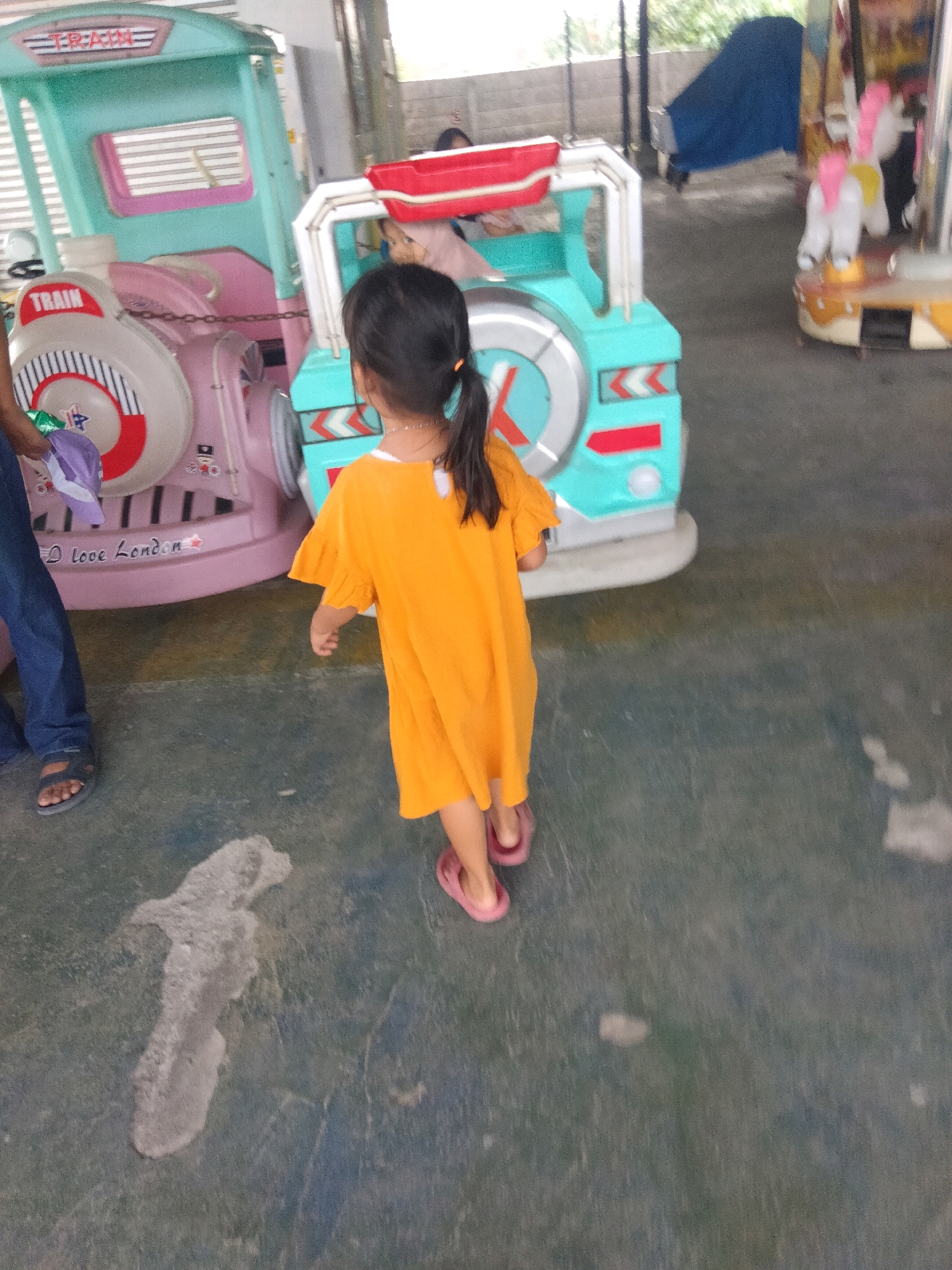 In Sukaasih, Indonesia, a young girl wearing an orange dress with ruffled sleeves walks away from the camera. She has dark hair pulled back in a ponytail and is wearing pink flip-flops. She is standing on a gray, textured ground surface with some light-colored stains and patches. To her left, a pink train-themed amusement ride is visible, with the word "TRAIN" printed on its side. Part of a person wearing blue jeans and dark sandals is visible on the far left. To the right of the girl, another amusement ride, possibly a carousel with white horses with pink manes and yellow tails, is partially visible. The background is out of focus but appears to be outdoors with some trees and structures.