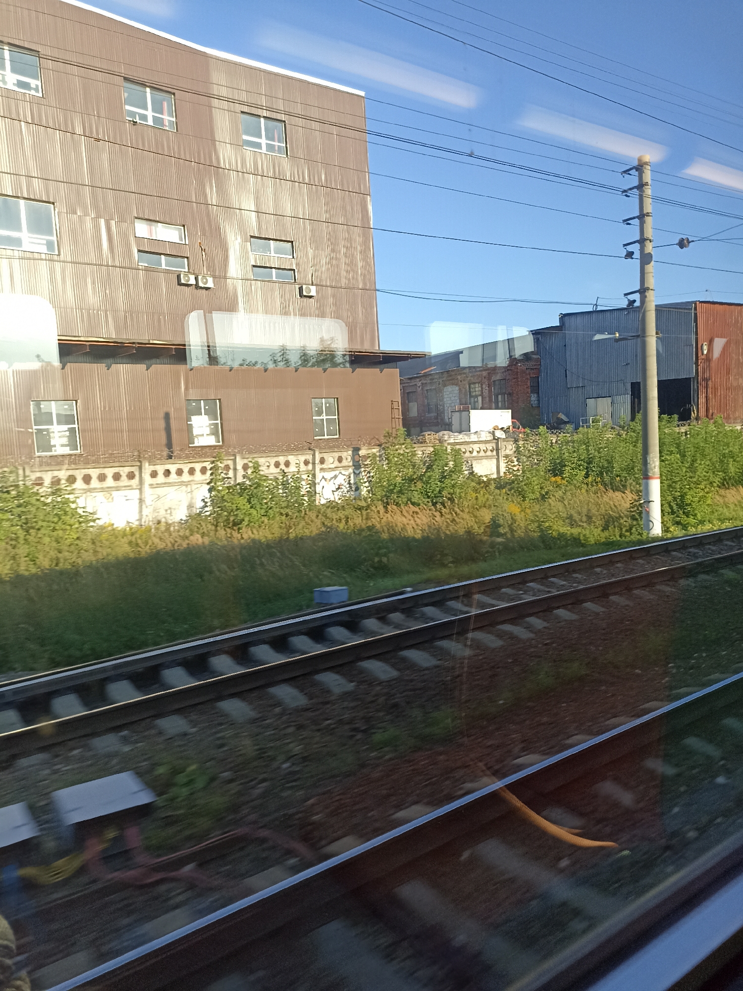 The image depicts an industrial area in Podolsk, Russia, viewed from a moving train. In the foreground, train tracks run diagonally across the frame, with the ballast and ties visible. Beyond the tracks, a grassy embankment leads up to a series of industrial buildings.
The most prominent building on the left is a multi-story structure with a corrugated metal facade and numerous windows. Several of these windows appear to have reflective blinds or curtains. Below this building, a lower structure with a similar corrugated metal exterior also features windows. Between these two buildings, an ornate concrete fence with hexagonal cutouts runs horizontally.
To the right of the main building, the landscape transitions to a collection of smaller industrial structures, including a brick building and a blue corrugated metal building. A utility pole with electrical wires stretches vertically along the right side of the frame. The sky above is clear and blue, with some reflections visible on the train's window. The overall scene suggests an active industrial zone during daylight hours.