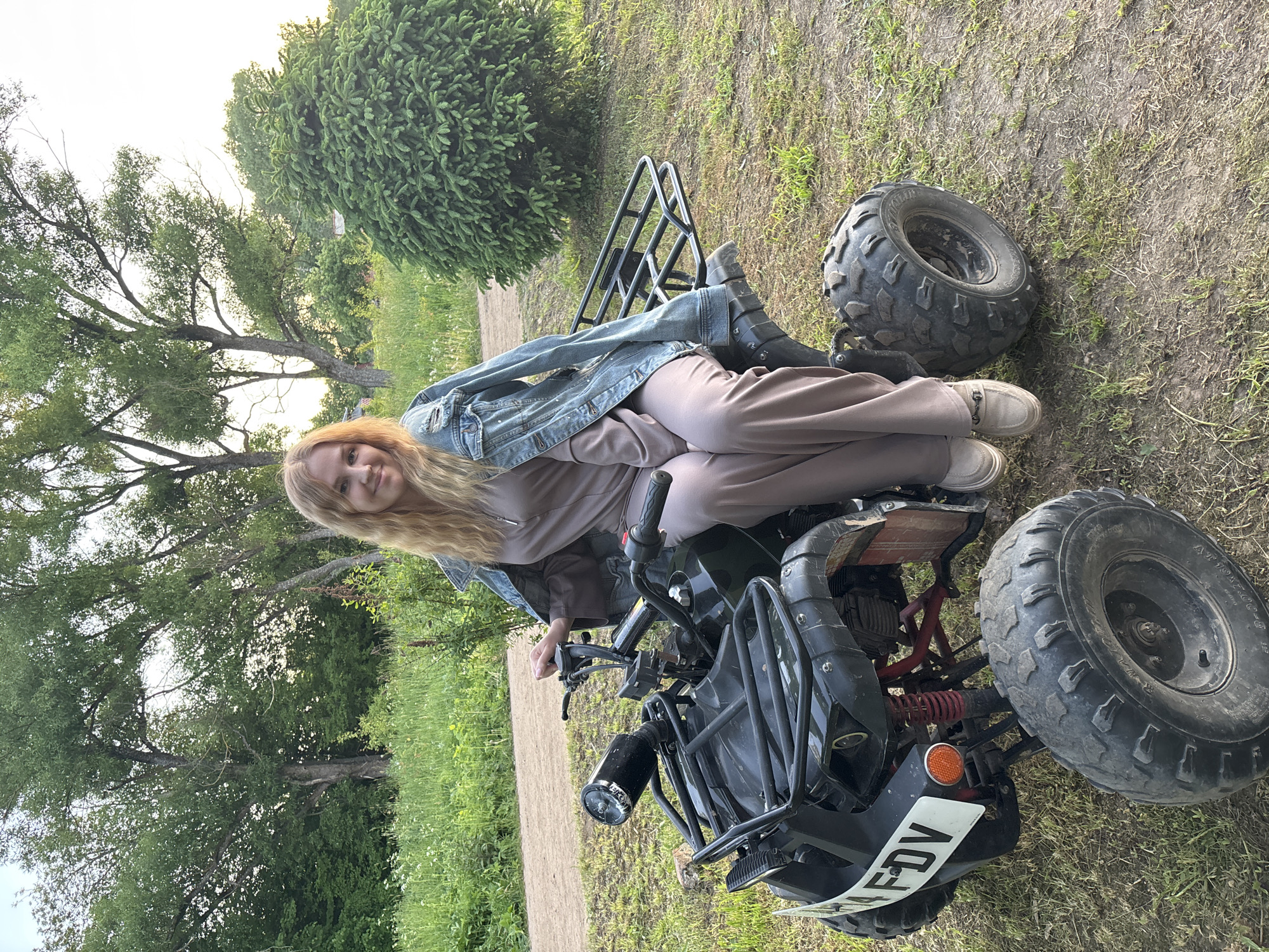 An outdoor scene in Rozhdestveno, Russia, depicts a young adult female seated on a dark-colored all-terrain vehicle (ATV). The female, with long blonde hair, smiles directly at the camera. She wears a light taupe-colored jumpsuit and a light blue distressed denim jacket adorned with white accents. Her left hand rests on the ATV's handlebar, and she wears light-colored loafers with metal detailing.
The ATV, possibly black or dark green, features prominent knobby tires, a black front rack with a cylindrical light source, and an orange circular reflector on the left front fender. A white license plate-like object, partially obscured, displays "FDV" on the front. Red suspension components are visible near the front wheel, and a black rack is present at the rear.
The environment is a natural setting, characterized by a dirt path traversing the mid-ground. Dense green grass and various wild plants cover the ground. Large deciduous trees with full green foliage form a backdrop, and a distinct, conical evergreen shrub is positioned on the right side of the frame. The lighting suggests late afternoon or early evening.