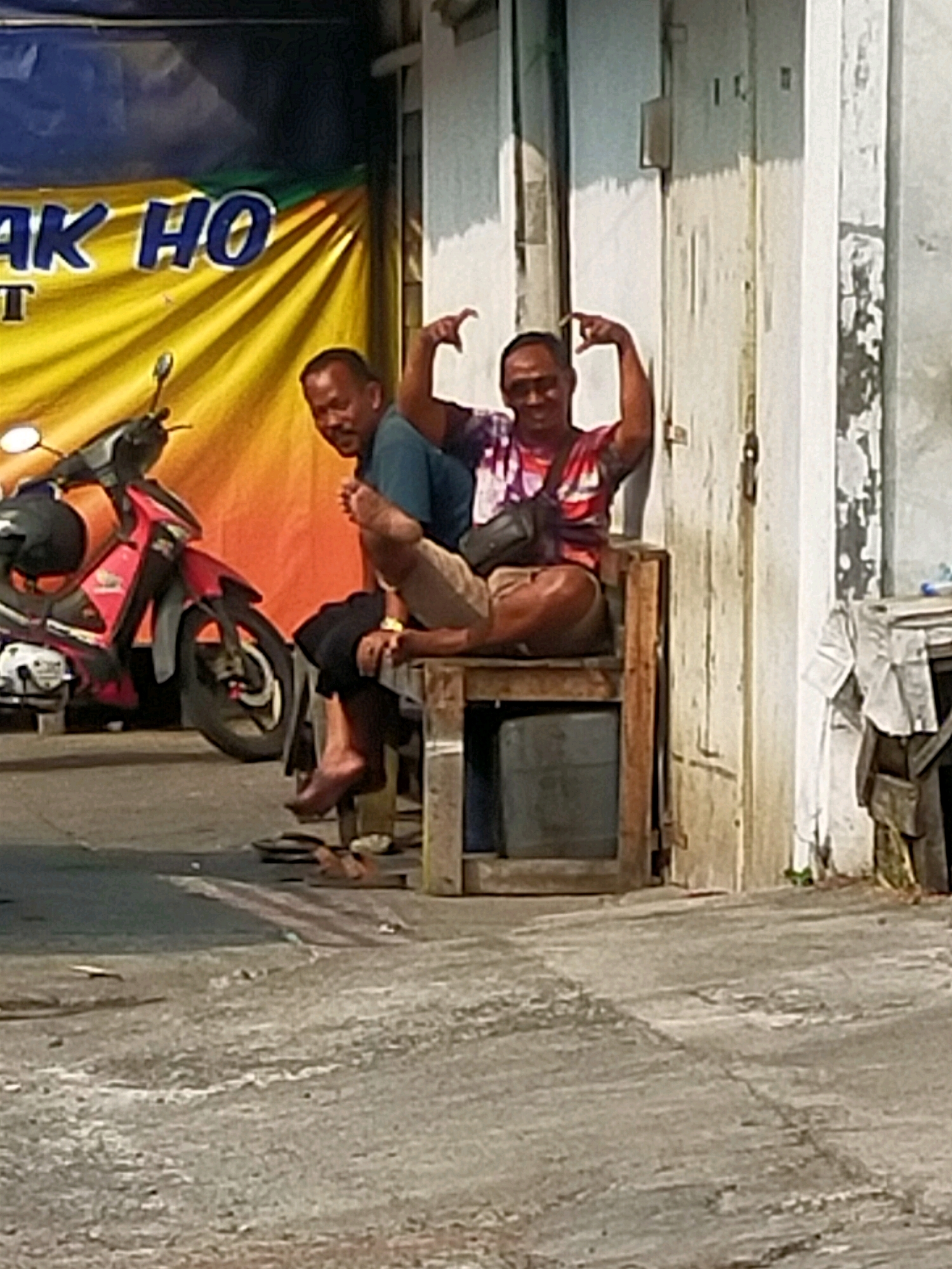 Two men are seated on a wooden bench in front of a building in what appears to be an urban setting in Surabaya, Indonesia. The man on the left, wearing a teal t-shirt, is looking towards the viewer with a slight smile and his legs crossed. The man on the right, wearing a tie-dye t-shirt and a black crossbody bag, is looking towards the viewer, striking a pose with his hands. A red and black motorcycle is parked behind the men. The background includes a yellow banner with blue text and a weathered building wall. The ground is paved with concrete, showing some cracks and debris.