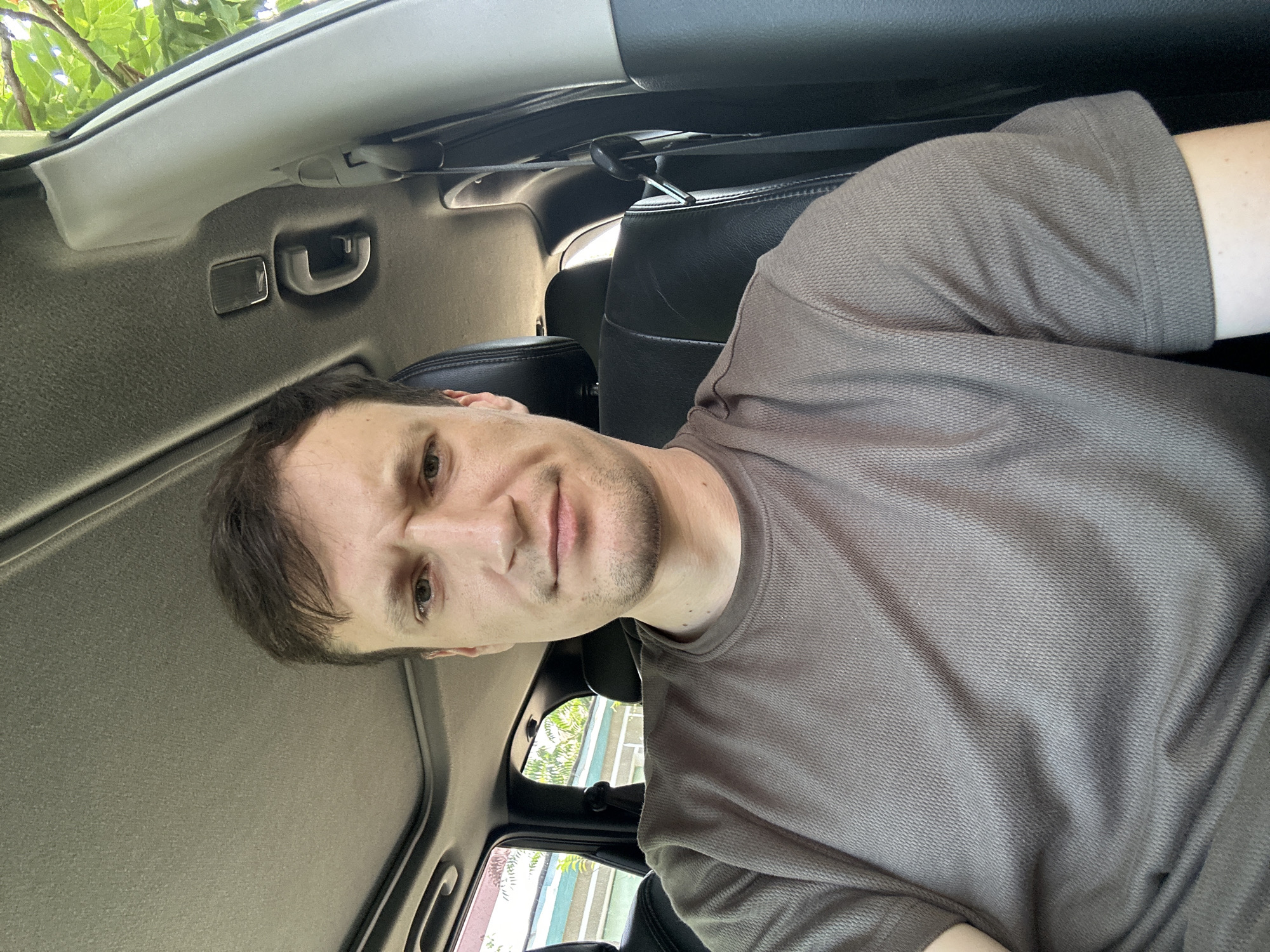 A medium shot shows a man with short brown hair and a light beard sitting in the driver's seat of a car. He is wearing a brown t-shirt with a textured weave. The man is looking directly at the camera with a serious expression, his eyebrows slightly furrowed. The interior of the car is visible, including the black leather headrest of the passenger seat behind him, the car's ceiling with a sunroof, and a portion of the passenger side door panel. Through the window behind him, a building with blue trim and greenery are visible, suggesting an outdoor, likely residential or commercial setting. The image is taken in the city of Makiv, Ukraine.
