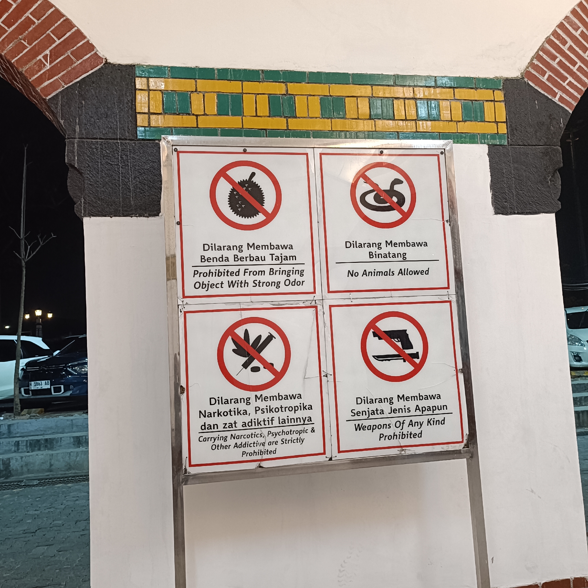 The image shows four prohibition signs mounted on a white wall, framed by a brick archway above. The signs are arranged in a 2x2 grid. The top left sign depicts a durian fruit with a red circle and slash, indicating it is prohibited. Below it, the text reads "Dilarang Membawa Benda Berbau Tajam" and "Prohibited From Bringing Object With Strong Odor". The top right sign shows a coiled snake with a red circle and slash, with text below reading "Dilarang Membawa Binatang" and "No Animals Allowed". The bottom left sign displays a syringe and some pills, also with a red circle and slash. The text below states "Dilarang Membawa Narkotika, Psikotropika dan zat adiktif lainnya" and "Carrying Narcotics, Psychotropic & Other Addictive are Strictly Prohibited". The bottom right sign shows a handgun and a knife crossed out with a red circle and slash, accompanied by the text "Dilarang Membawa Senjata Jenis Apapun" and "Weapons Of Any Kind Prohibited". The environment appears to be outdoors, with a dark background suggestive of nighttime. A dark-colored car with a license plate visible (H 1863 AB) is parked to the left of the signs. The architecture suggests an archway or entrance, possibly in a public space. The overall context implies a security or facility regulation within the city of Semarang, Indonesia, given the Indonesian text on the signs.