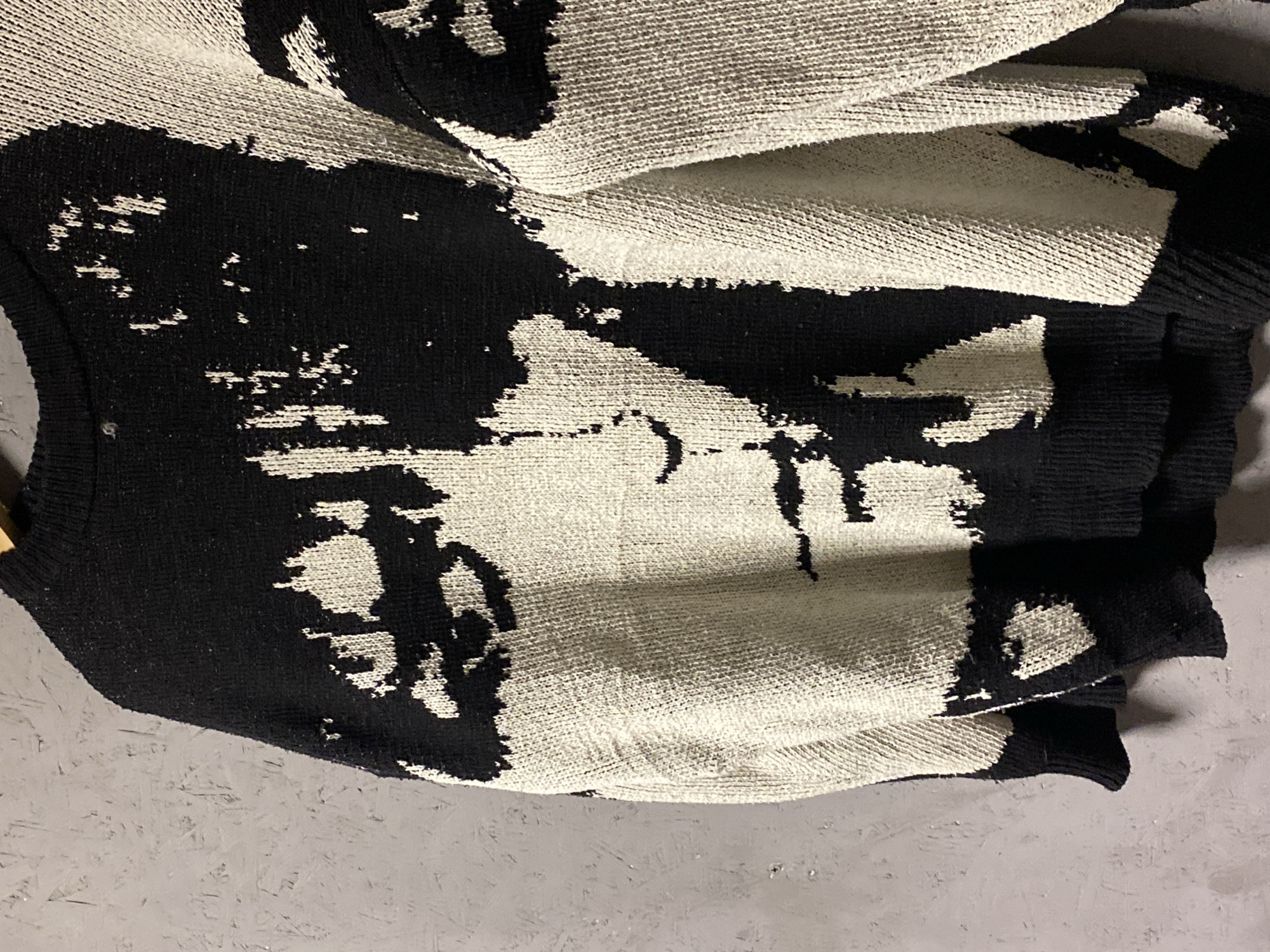 A close-up, medium shot shows a black and cream-colored knit sweater with a large, abstract portrait design. The portrait appears to be a face rendered in bold black and off-white blocks. The sweater is draped over a surface, possibly a clothes hanger, against a textured gray wall. The lighting is dim, creating shadows that emphasize the texture of the knit. No specific location in Krasnoyarsk, Russia, can be determined from the image.