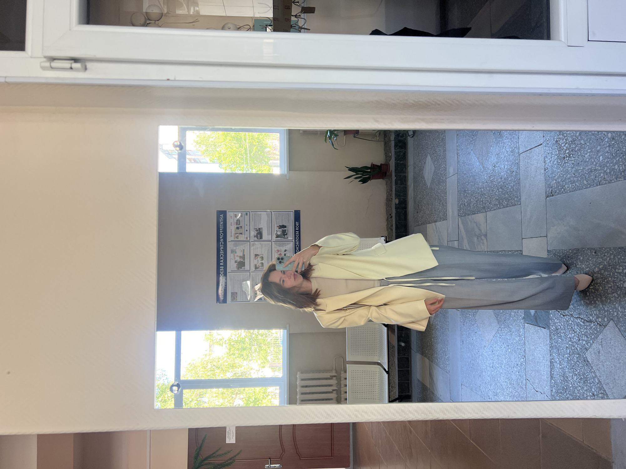 A young woman is taking a selfie in a mirror. She is standing in a hallway with a tiled floor and windows on one side. She is wearing a pale yellow blazer over a tan top and light blue wide-leg pants. She has medium-length brown hair and is holding a phone in her right hand. In the reflection, behind her, there are two rows of white chairs, a radiator, and a notice board on the wall. The environment appears to be indoors, possibly in a school or administrative building, given the presence of chairs and a notice board. The location is identified as the city of Chaikovsky, Russia, though no specific landmarks are visible to confirm this.