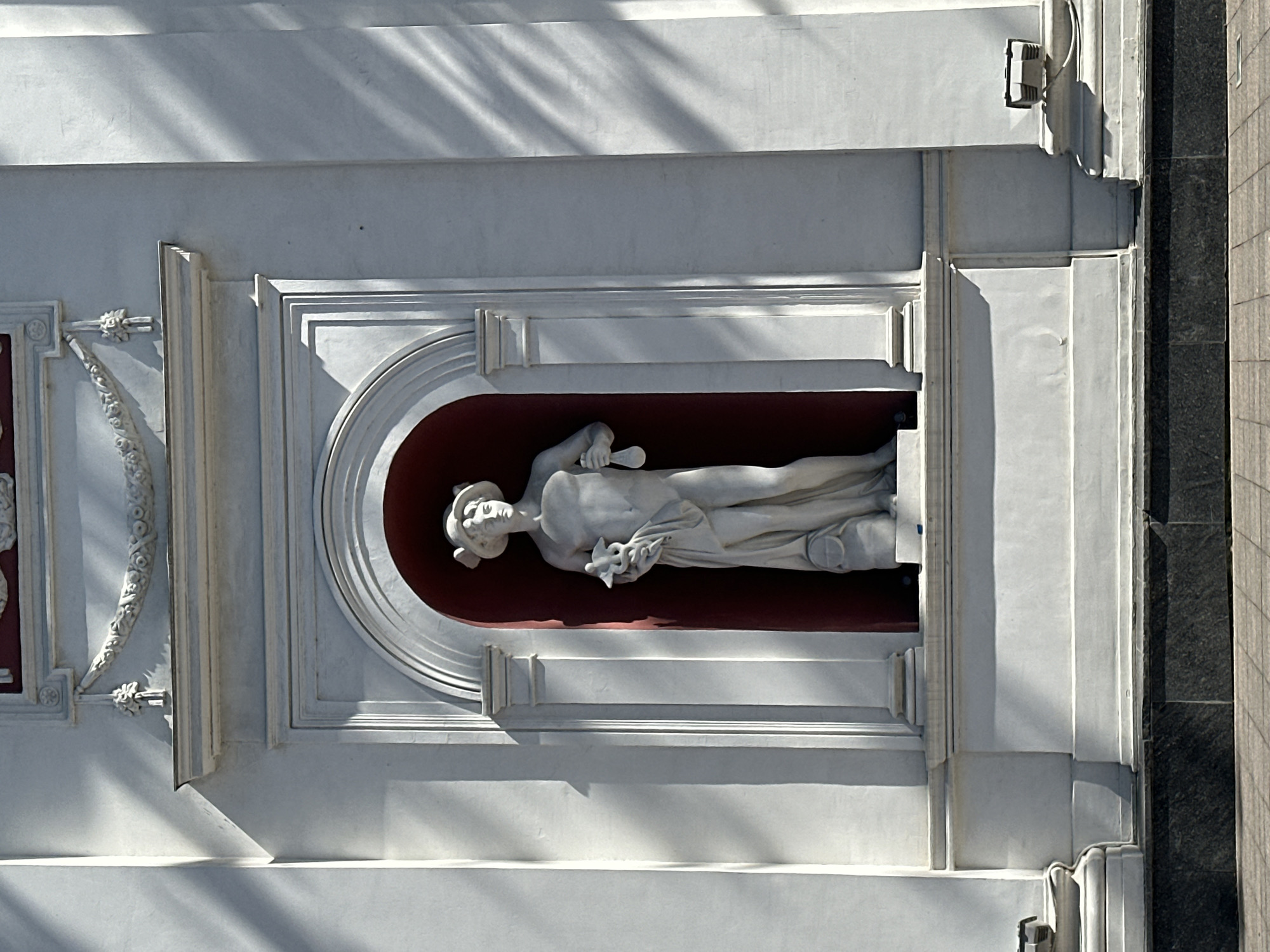 A full shot shows a stone sculpture of a man framed in white, set into the white facade of a building in Unknown, Unknown. The statue is in an alcove with a deep red backing. The figure wears a head covering, a robe, and carries something in its left hand and a staff in its right. The background of the building is plain white with classical decorations. The shadow lines of the building cut diagonally across the facade. A wall of marble-like brick sits on the right side of the frame.