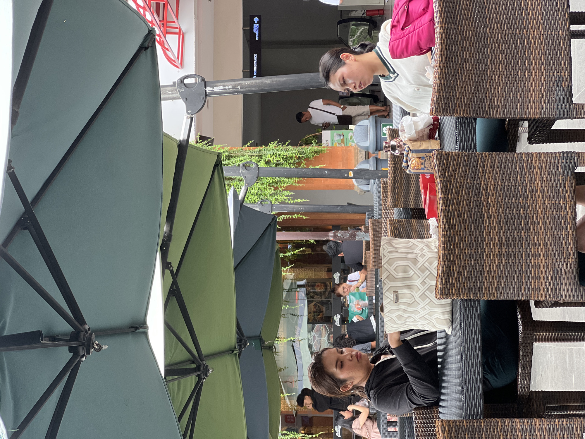 The image shows a street-level view of an outdoor cafe in Kuta, Indonesia. Several tables with woven chairs are positioned under large green patio umbrellas. People are seated at the tables, some engaged in conversation. In the background, pedestrians walk along the sidewalk and greenery is visible. Signage is partially visible.