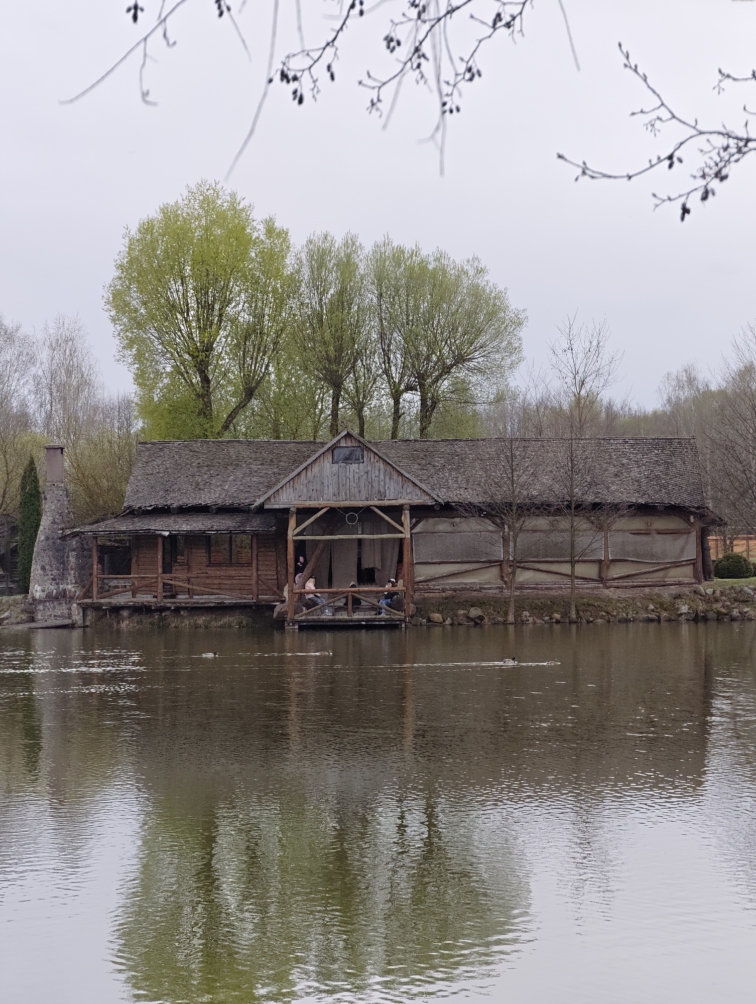 The image depicts a wooden structure situated on a body of water, likely a lake or pond, in Капцёўскі сельскі Савет, Belarus. The structure has a dark, shingled roof and is constructed of logs and timber. A chimney is visible on one side. The building is positioned partially over the water, with a section appearing to be a covered deck or patio. Two people are seated on the deck. The water reflects the structure and the surrounding trees. Trees with green leaves are in the background, along with bare trees. The sky is overcast. A tree branch with buds is in the top of the image. Several ducks are swimming in the water.