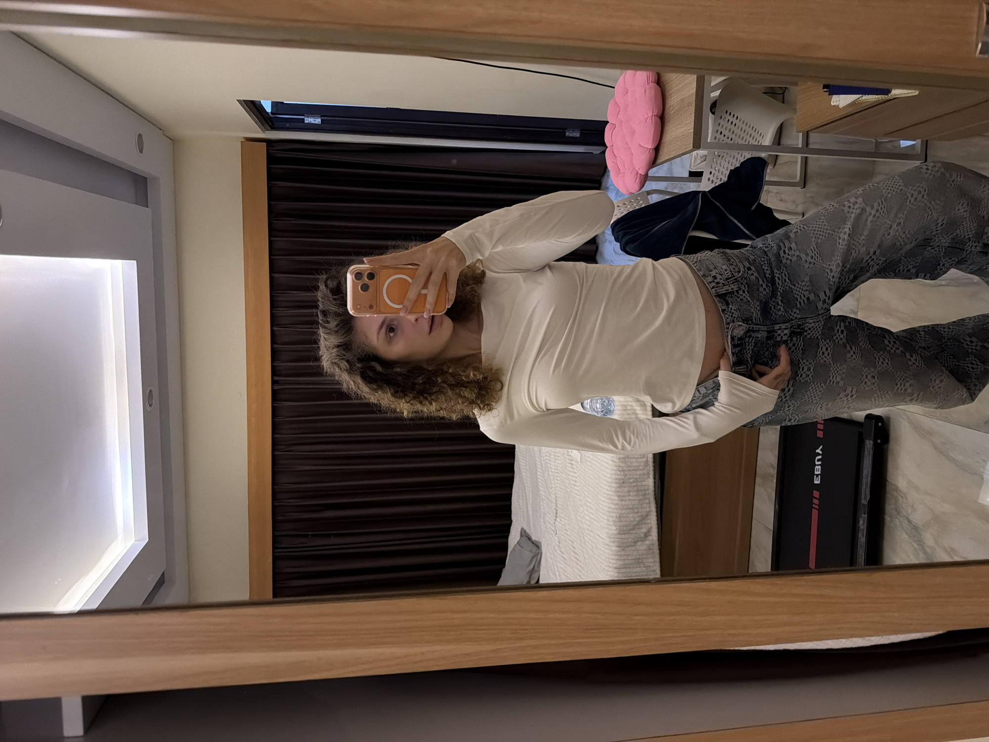 A person is taking a mirror selfie in what appears to be a hotel room in North Kuta, Indonesia. The individual is wearing a white long-sleeved top and patterned jeans, holding an orange iPhone in their right hand. The reflection reveals a room with a bed, dark curtains, a desk with a pink cushion on it, and a white plastic chair. A black foldable treadmill is visible in the lower right corner. The room's interior design is modern, with a lighted ceiling feature and wooden furniture.