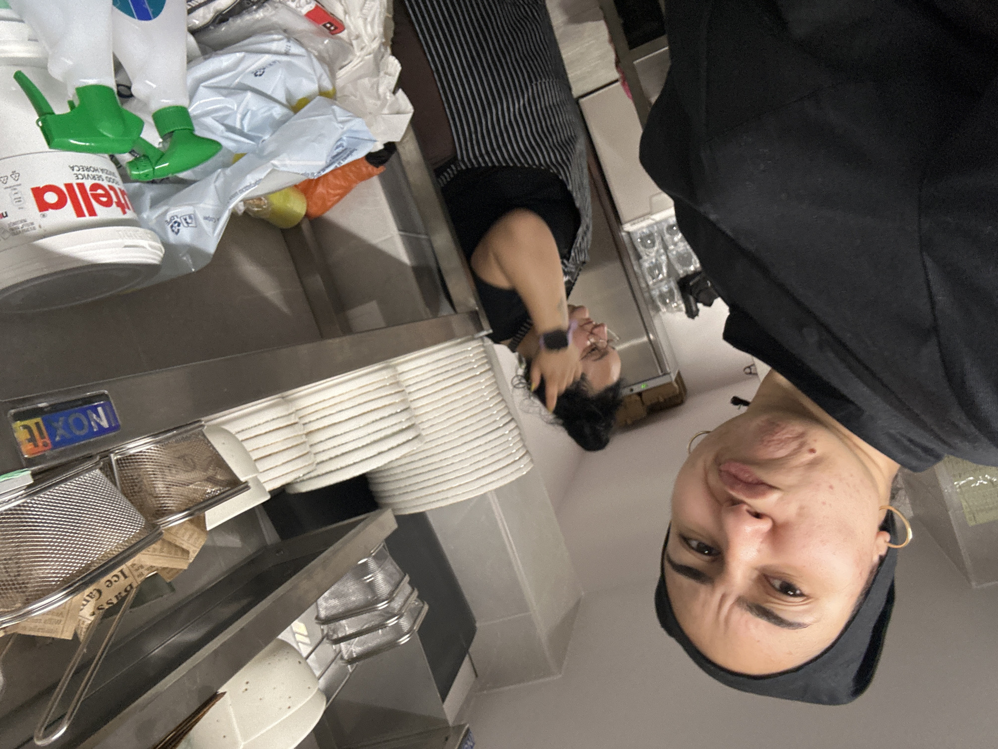 The scene appears to be the interior of a commercial kitchen, potentially in a restaurant or food preparation area. Two individuals are present. One person is positioned in the foreground, facing the camera. They have dark hair, are wearing a black head covering, and a black shirt. Gold hoop earrings are visible. The second person is further back, wearing a black and white striped apron. A stack of white plates and a metal shelving unit containing various items, including a metal strainer and what appears to be newspaper, are also present. A container of Nutella is visible near the plates. The lighting is bright, suggesting artificial illumination within the kitchen environment. There is no definitive information in the image to confirm the location of Burgas, Bulgaria.