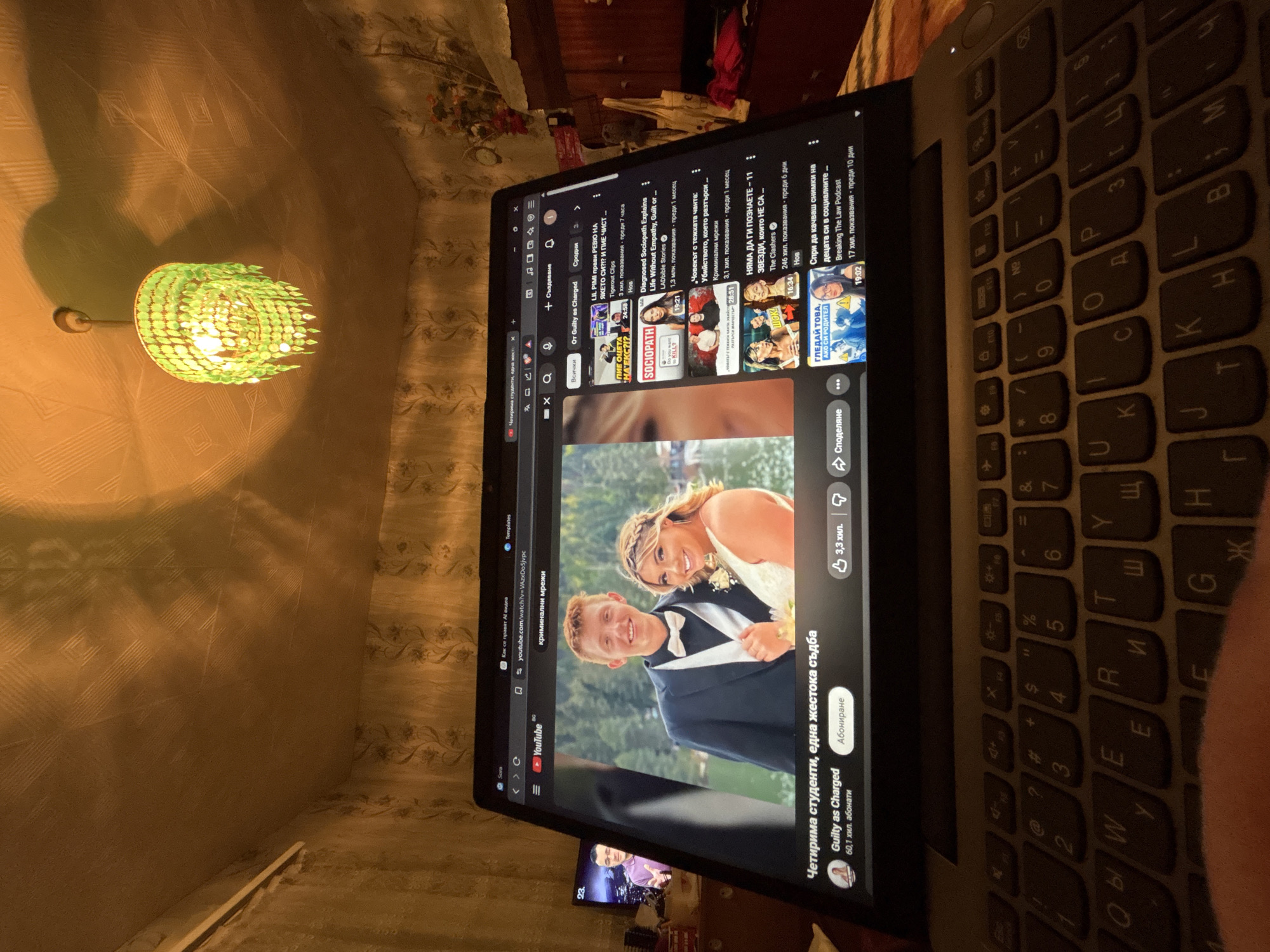 A laptop is on a table in a room. The screen shows a YouTube video titled "Четирима студенти, една жестока съдба" (Four students, a cruel fate) by the channel "Guilty as Charged." The video thumbnail shows a smiling couple. In the background, there is a lamp with a decorative green lampshade, and a TV showing some kind of broadcast. The room appears to be a bedroom or living room, but the location in the city of Dobrovnitsa, Bulgaria, cannot be verified from this image. There is no discernible interaction between people.
