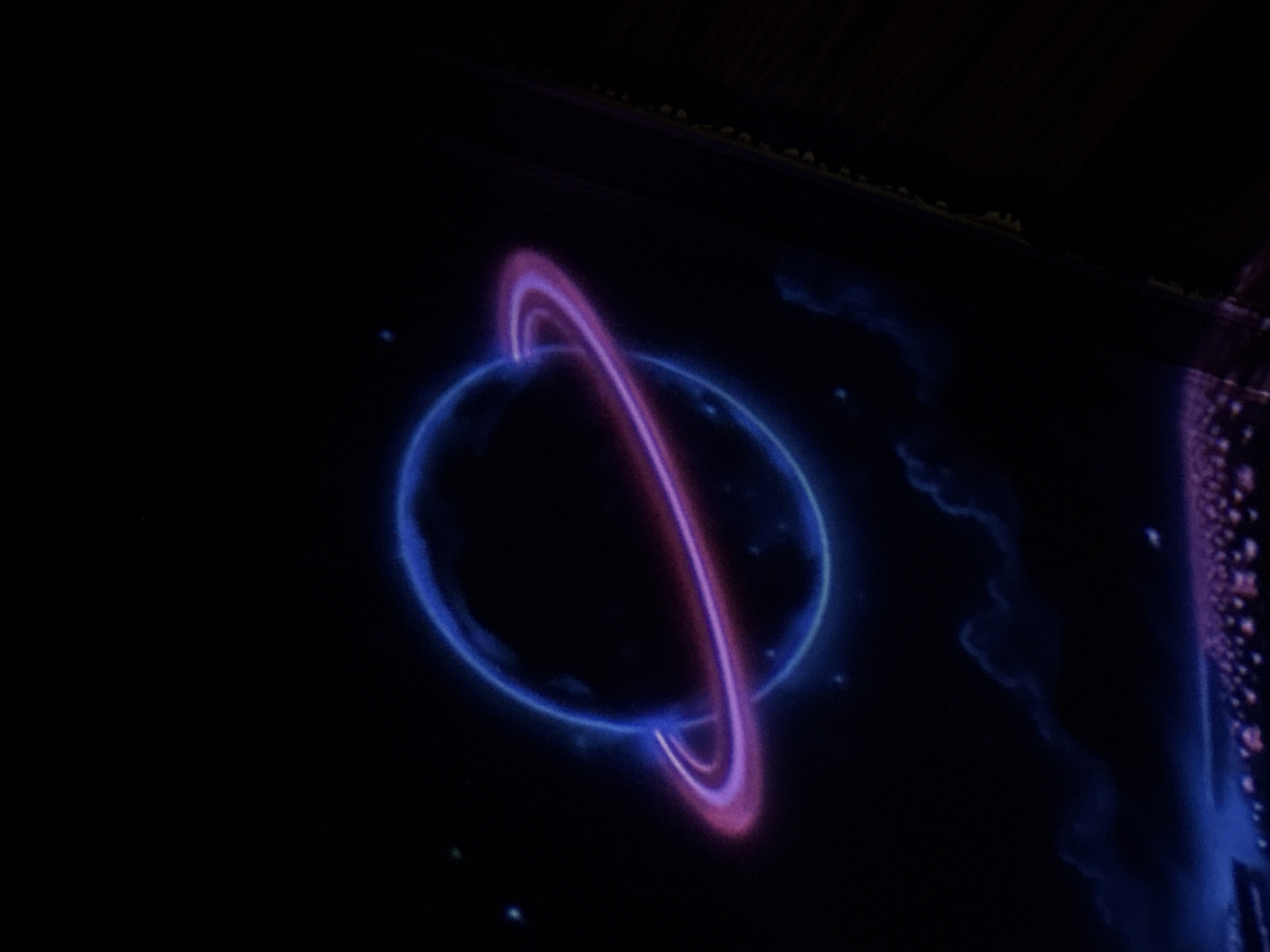 The image depicts a neon-style depiction of a ringed planet against a black background, suggesting a cosmic or otherworldly theme. Below the planet, a stylized representation of clouds and a city landscape with glowing lights is visible. The city appears to be densely populated with structures and illuminated areas. The overall aesthetic is dark, with vibrant neon colors dominating the planet and its rings, and subtle glows emanating from the city. There are no people or specific actions depicted, only a static scene. The image is an artistic representation and does not contain identifiable landmarks or features that would place it in a specific city like Tbilisi, Georgia.