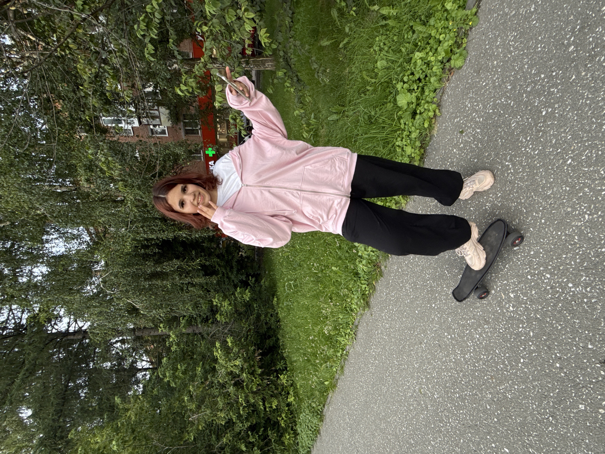A young woman with reddish-brown hair stands on a skateboard on a paved pathway in Yekaterinburg, Russia. She is wearing a light pink zip-up hoodie over a white t-shirt and loose black pants, with light beige athletic shoes. The skateboard is black and appears to be a penny board. The pathway is bordered by a strip of green grass and foliage, with trees in the background. Behind the trees, a multi-story brick building with windows is visible. The woman is smiling and holding a smartphone in her right hand, with her left hand near her face. The overall environment suggests an outdoor urban setting.