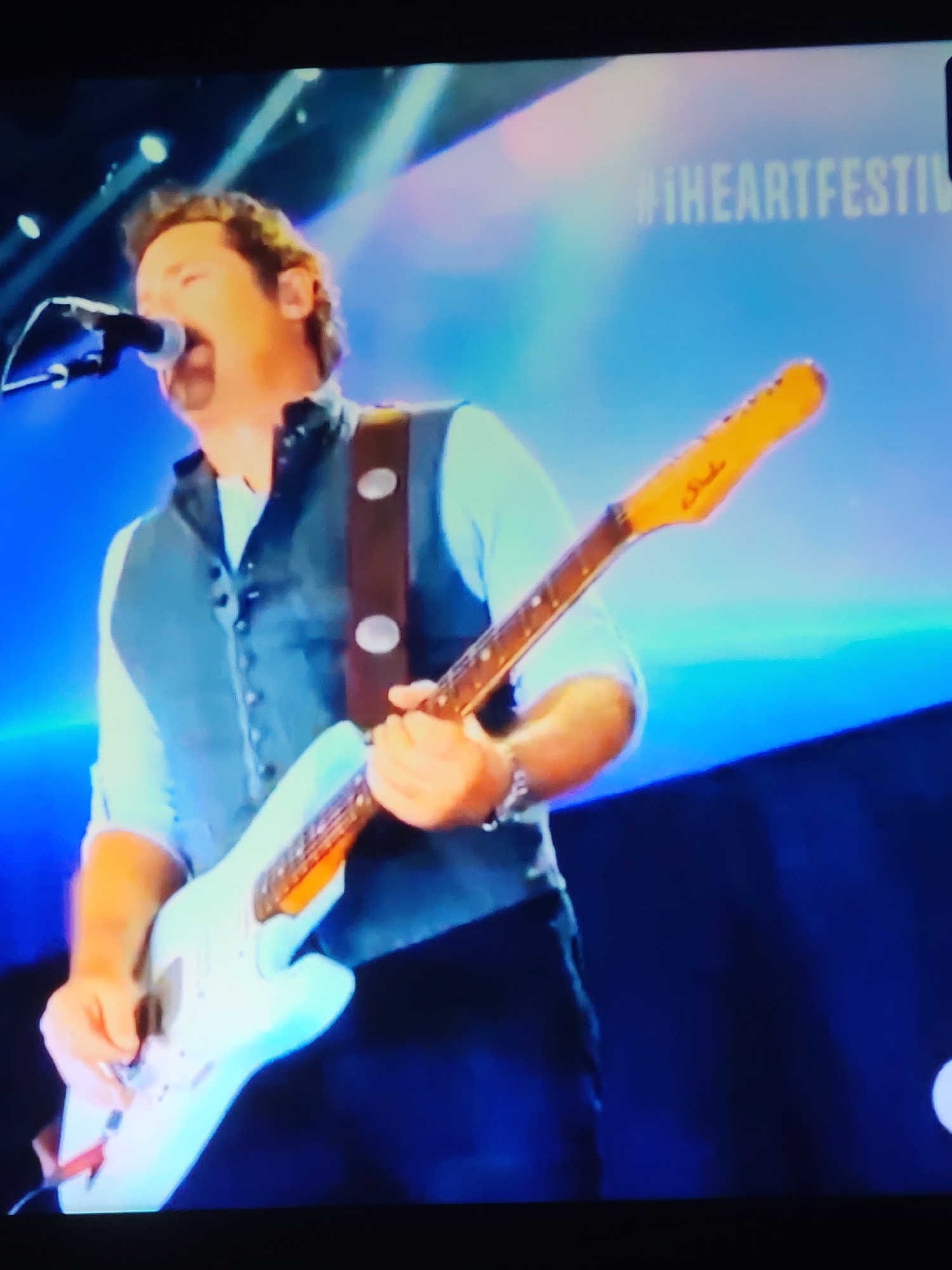 A man is singing into a microphone while playing a light blue electric guitar on a stage. He is wearing a white shirt, a dark gray vest with buttons, and a brown strap with two circular embellishments. The background is a bright blue with some soft, out-of-focus lights. Text that appears to read "#IHEARTFESTIVAL" is visible in the upper right corner of the background. The image is blurry, suggesting it was taken from a distance or is a low-resolution capture. There is no information in the image to suggest a location in Vladimir, Russia.