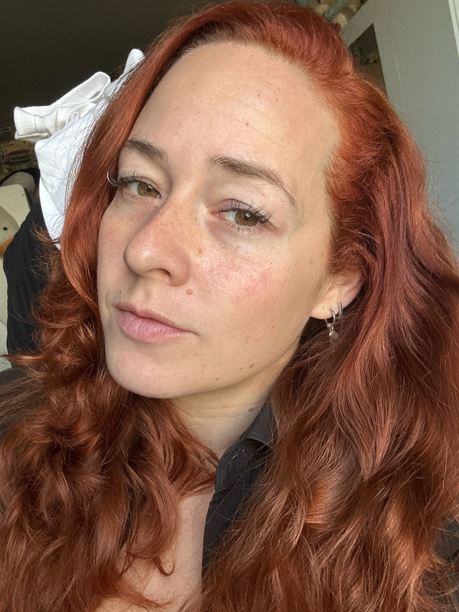 A medium-shot, head-and-shoulders portrait of a woman with auburn hair, freckles, and green eyes. She is wearing a black collared shirt and silver hoop earrings. Her hair is wavy and falls past her shoulders. The background is out of focus and appears to be an indoor setting with natural light. There is no discernible Moscow location.