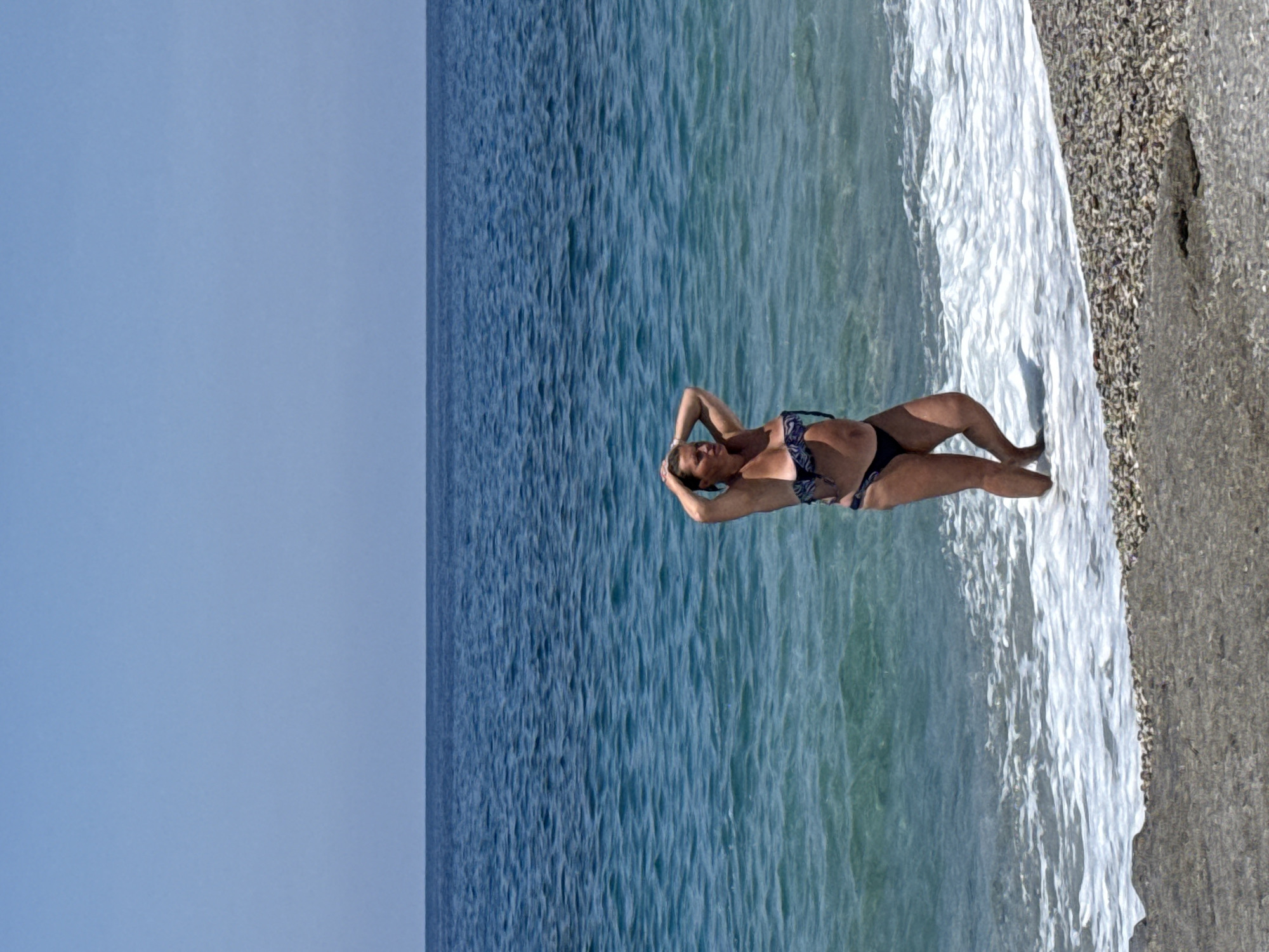 An outdoor, eye-level, full-shot photograph depicts an adult female in a two-piece bikini standing at the water's edge on a pebbled beach in Alanya, Turkey. The woman, positioned slightly right of the frame's center, has light skin and her dark hair is pulled back, with her hands clasped behind her head. She wears a dark blue patterned bikini top and a solid dark bikini bottom. Her right leg is slightly lifted, with her left foot planted in the shallow, clear turquoise water, which extends to her mid-calves. Gentle white foam from breaking waves is visible around her feet and along the shoreline. The sea, a deep blue further out, meets a clear, light blue, cloudless sky at a distinct, flat horizon line. The immediate foreground displays the wet, grey and brown pebbled beach.