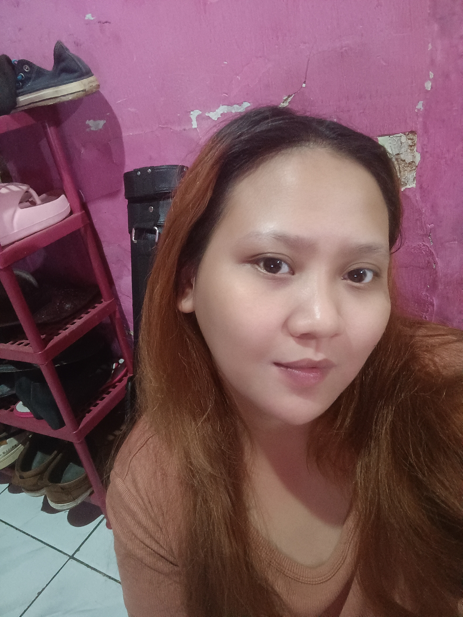 In a room with a pink, peeling wall, a woman with long, reddish-brown hair takes a selfie. She is wearing a rust-colored, ribbed t-shirt and her face is partially visible. To her left, a red plastic shelf unit holds several pairs of shoes, including pink slides and brown loafers. A black, rectangular object, possibly a bag, leans against the wall behind her. The floor is tiled in white squares with grey grout lines. The scene appears to be an indoor domestic setting within a residential area of Depok, Indonesia.