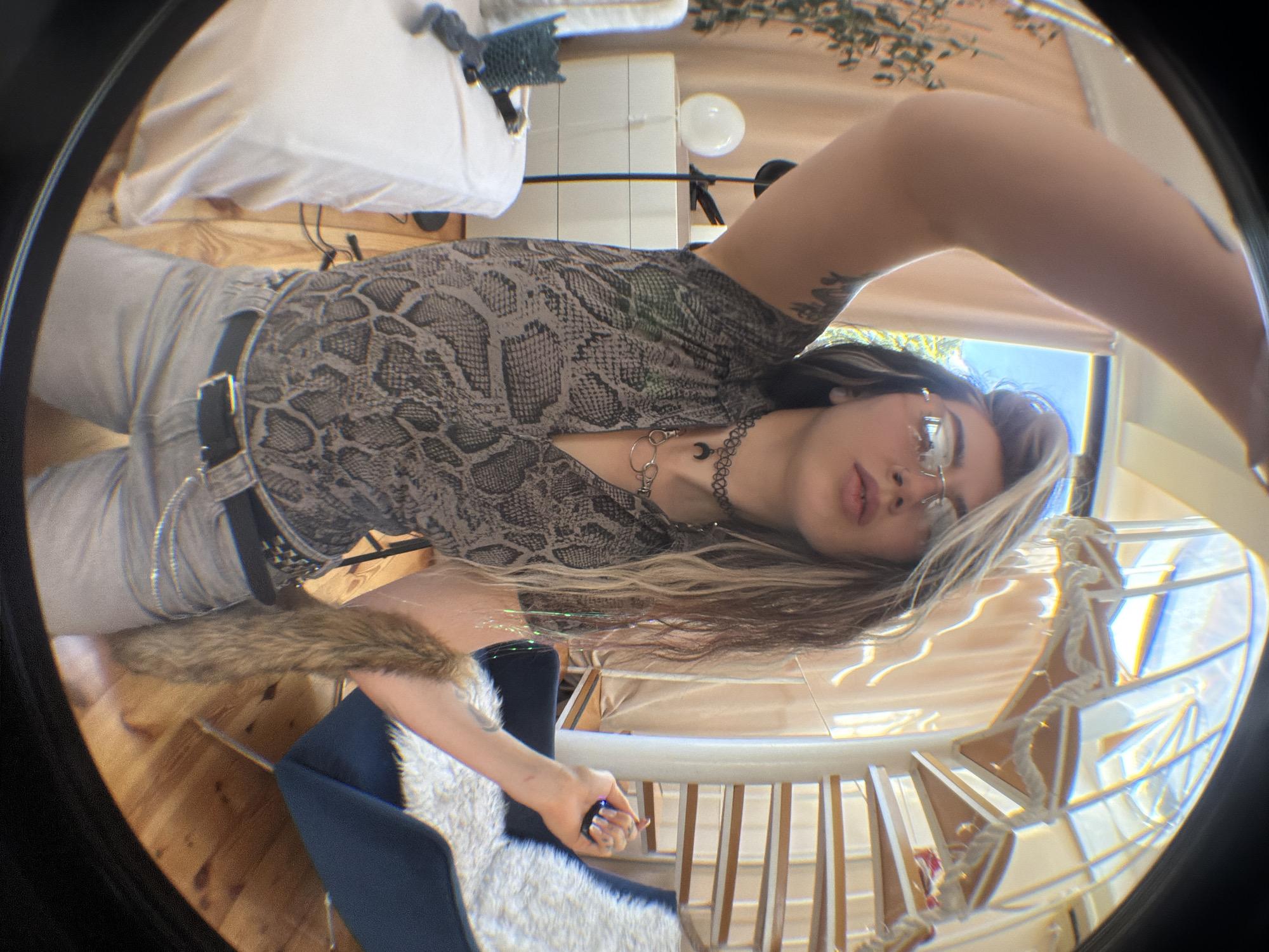 A wide-angle, fisheye lens photograph captures a young individual standing indoors, posing for a selfie. The subject, positioned centrally, has long, multi-toned hair with dark and blonde streaks, wears rimless rectangular glasses, a sleeveless grey and black snakeskin-patterned top, light-colored trousers, a black belt with a silver buckle, and two necklaces (a black choker with a crescent moon charm and a silver-toned chain). Visible tattoos are on the subject's raised and bent left arm. The right hand holds a small, dark object emitting a blue light. A brown, furry accessory is draped over the right forearm. The background reveals a brightly lit interior space, likely a modern apartment or hotel room in Budva, Montenegro. Features include a white spiral staircase with light wood steps and rope detailing on the right, a white-covered bed or sofa with a small dark container and grey fabric item on the left, and a white three-drawer unit with a round white lamp behind the subject's left arm. A dark blue armchair with a white faux fur throw is present on the right. The floor is light-colored wood, and beige curtains are visible in the background.