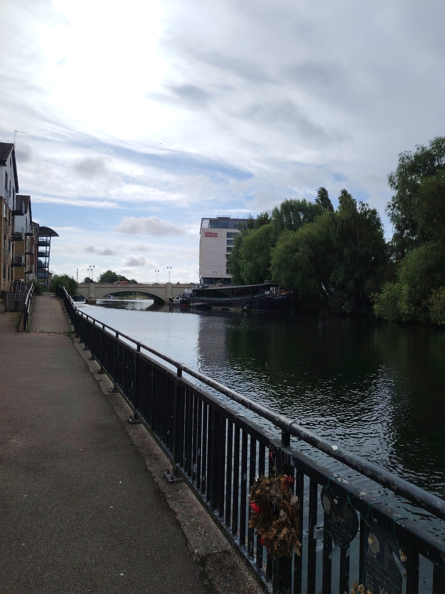 The image depicts a riverside scene in Peterborough, United Kingdom, specifically along the River Nene embankment, looking southeast towards the Town Bridge and the Hilton Garden Inn hotel.
In the foreground, an asphalt path occupies the left side, paralleling the river. A black metal railing separates the path from the dark, rippling water of the River Nene, which fills the right side of the frame. Attached to the railing in the lower right corner is a wreath made of dried brown leaves with subtle red accents, alongside several dark, heart-shaped plaques or locks.
In the midground, a light-colored, multi-arched stone bridge (the Town Bridge) spans the river. A red vehicle is visible crossing the bridge, which is lined with lampposts. To the right of the bridge, on the opposite bank, stands the white Hilton Garden Inn hotel building with its red logo prominent. Further along the opposite bank to the right, a dark, flat-topped boat or barge, labeled "LLENROB R," is moored, partially obscured by a dense cluster of green deciduous trees. Residential buildings with balconies line the left bank in the middle distance.
The background features a cloudy sky, predominantly white and light grey, with some darker patches indicating an overcast but bright day. No people are visible.