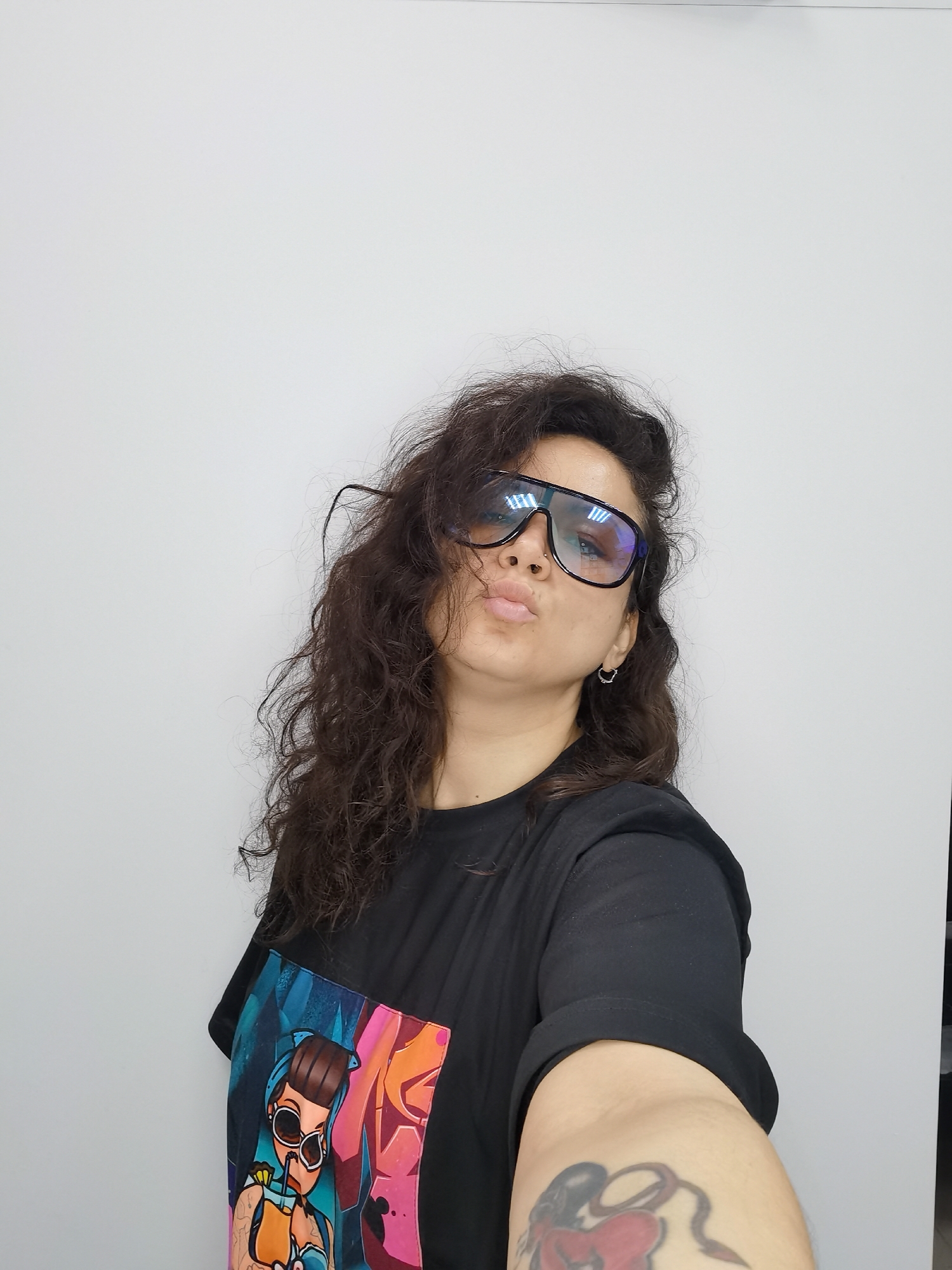 A woman with long, dark, curly hair and a black t-shirt with a graphic design is posing for a selfie against a plain white background. She is wearing large, dark-rimmed glasses and has her lips pursed. A silver hoop earring is visible in her right ear. Her left arm is extended towards the camera, and a tattoo of a red heart with a black ribbon is visible on her upper arm. The image appears to be taken indoors, with consistent lighting and no visible environmental details to suggest a specific location in Chaikovsky, Russia.