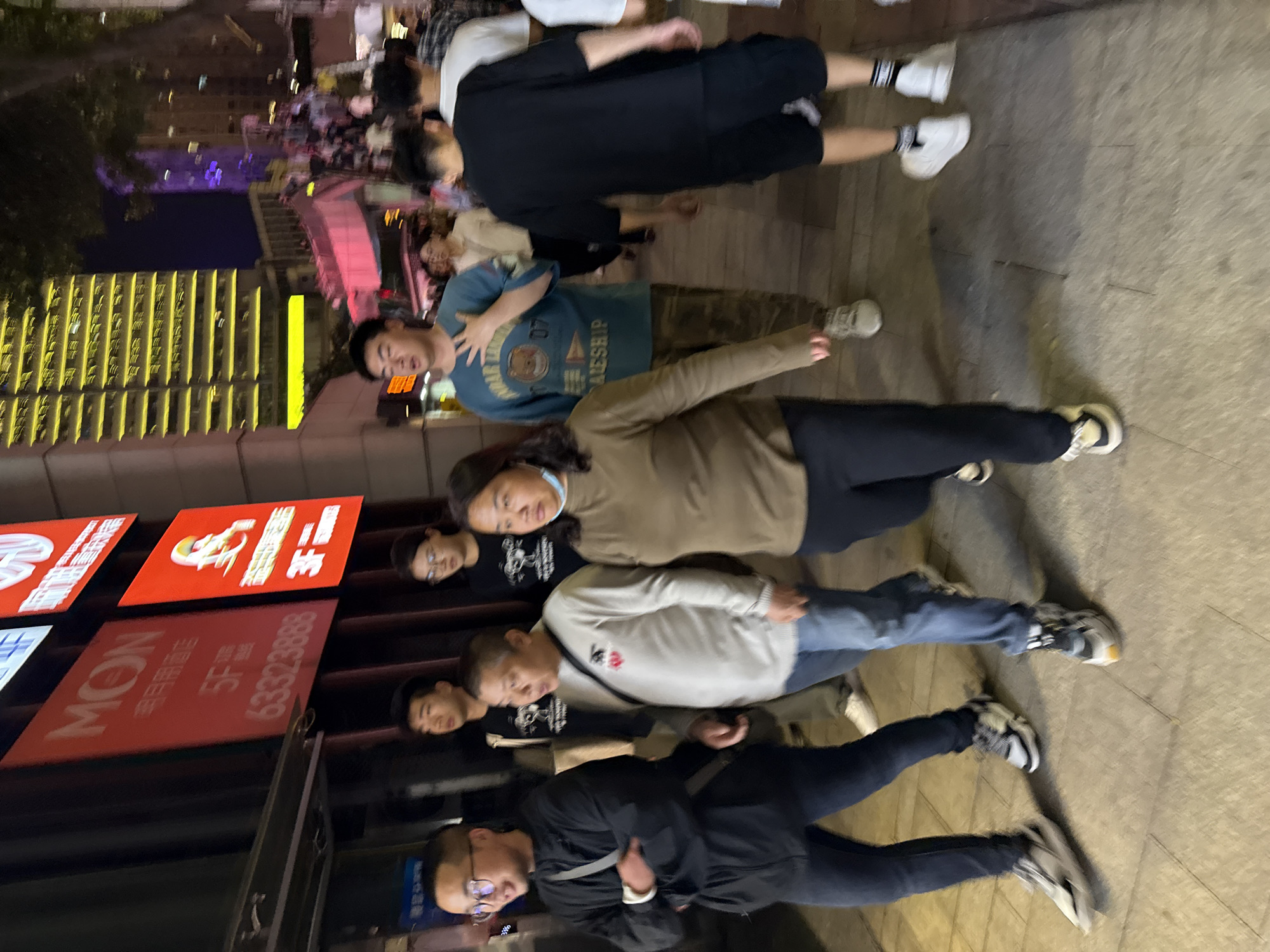 This media file captures a nighttime street scene in an urban area, likely Yuzhong District, China, showing a group of East Asian individuals standing on a paved sidewalk. The image is oriented vertically, with subjects arranged across the frame from left to right.
Approximately eight people are visible, dressed in casual attire including jeans, t-shirts, sweaters, and sneakers. Their expressions are generally neutral or contemplative. One person near the center wears a light blue t-shirt with the number "07" and the word "BALESHIP" printed on it, along with a bear graphic. Other clothing includes dark jackets, a light brown long-sleeved top, and a white sweater with a small red embroidered detail. Most individuals are wearing sneakers in various light and dark shades.
The background features brightly lit multi-story buildings, typical of a city environment at night. Yellow lights illuminate windows on one building, while another structure in the distance glows with purple light. Artificial street lighting is also present.
On the far left, several large, vertically oriented signs are visible. One red sign prominently displays "MON" in English, accompanied by Chinese characters, possibly indicating "酒店" (hotel), and the phone number "63323888". Another red sign above it features white text and a logo that resembles the KFC Colonel Sanders figure, alongside more Chinese characters.
The scene depicts people gathered or pausing on the sidewalk. There isn't a specific event taking place beyond general urban activity. The time of day is clearly night, characterized by the dark sky and pervasive artificial lighting. The weather appears dry. Some motion blur is noticeable, particularly in the background elements and the feet of some individuals, suggesting either movement by the photographer or subjects, or a longer exposure time in low light.