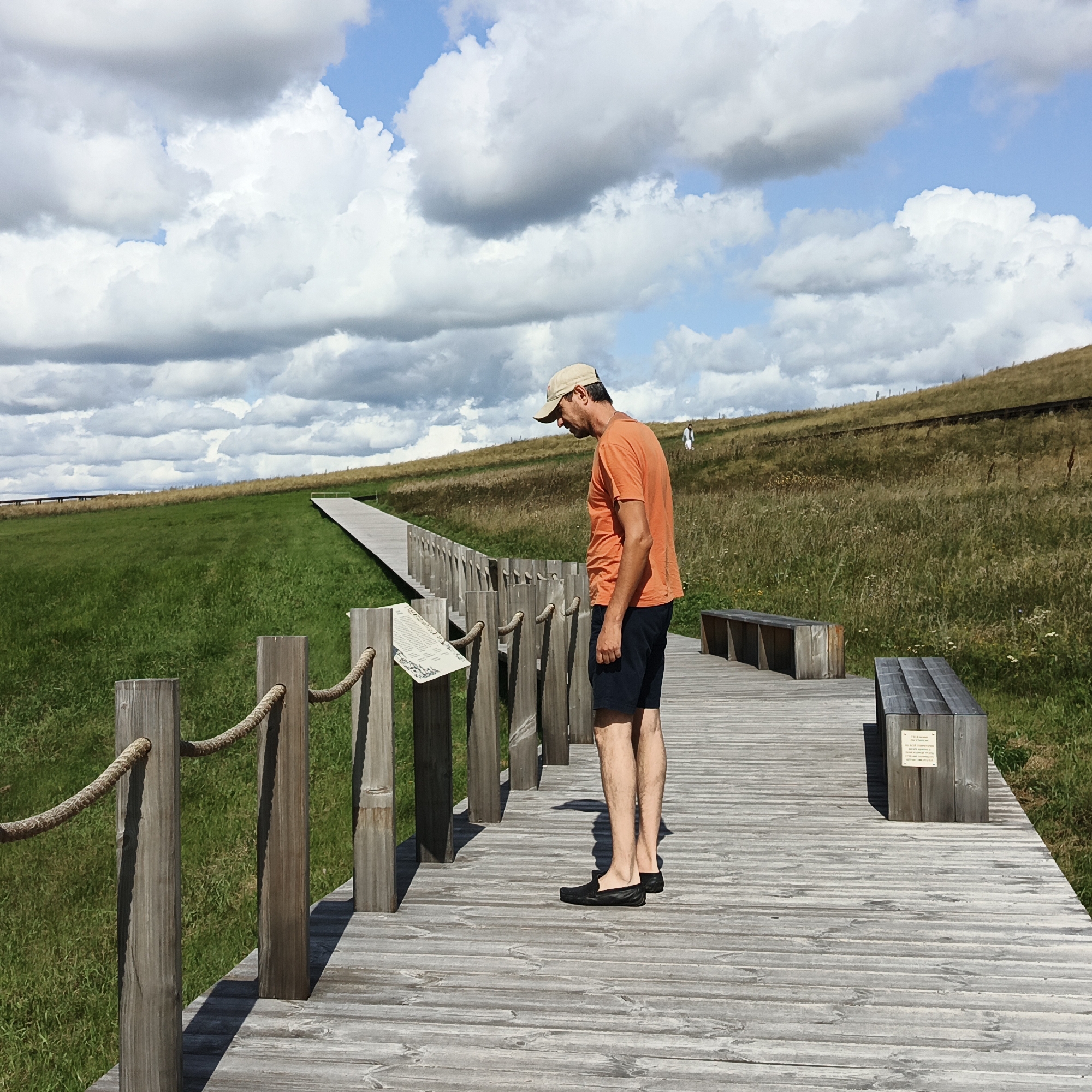 In Verkhnie Termy, Russia, a man wearing a beige baseball cap, an orange t-shirt, and dark shorts stands on a wooden boardwalk. He is facing away from the camera, looking down at an informational sign mounted on wooden posts. The boardwalk stretches into the distance, bordered by a rope railing and lush green grass. In the background, a gentle hill covered in dry grass rises under a partly cloudy sky. Further up the hill, another person in white clothing can be seen. Two wooden benches are placed along the boardwalk. The scene appears to be an outdoor recreational area.